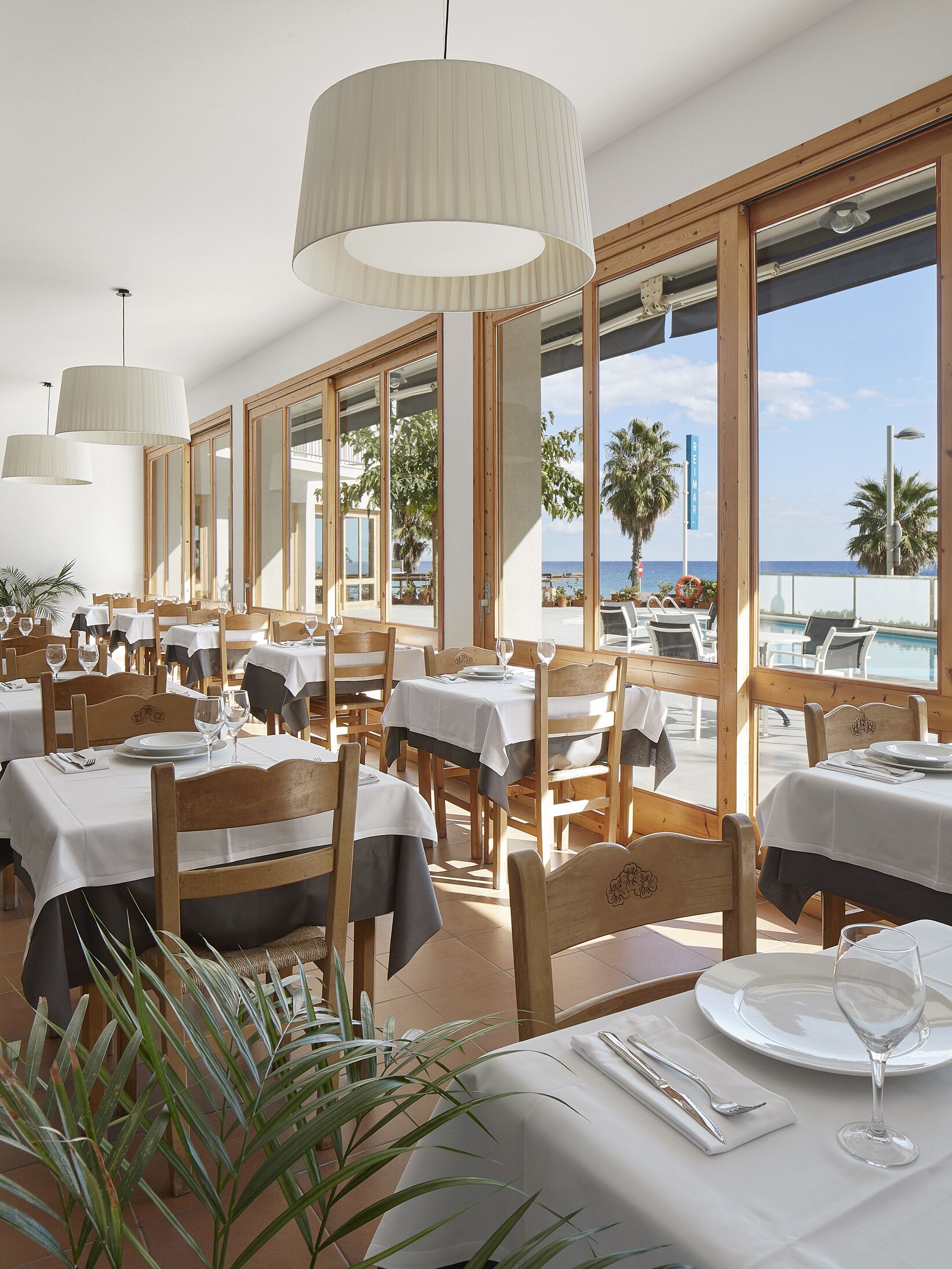 Un restaurante con varias mesas y sillas, ventanas amplias con vista al mar y lámparas colgantes.