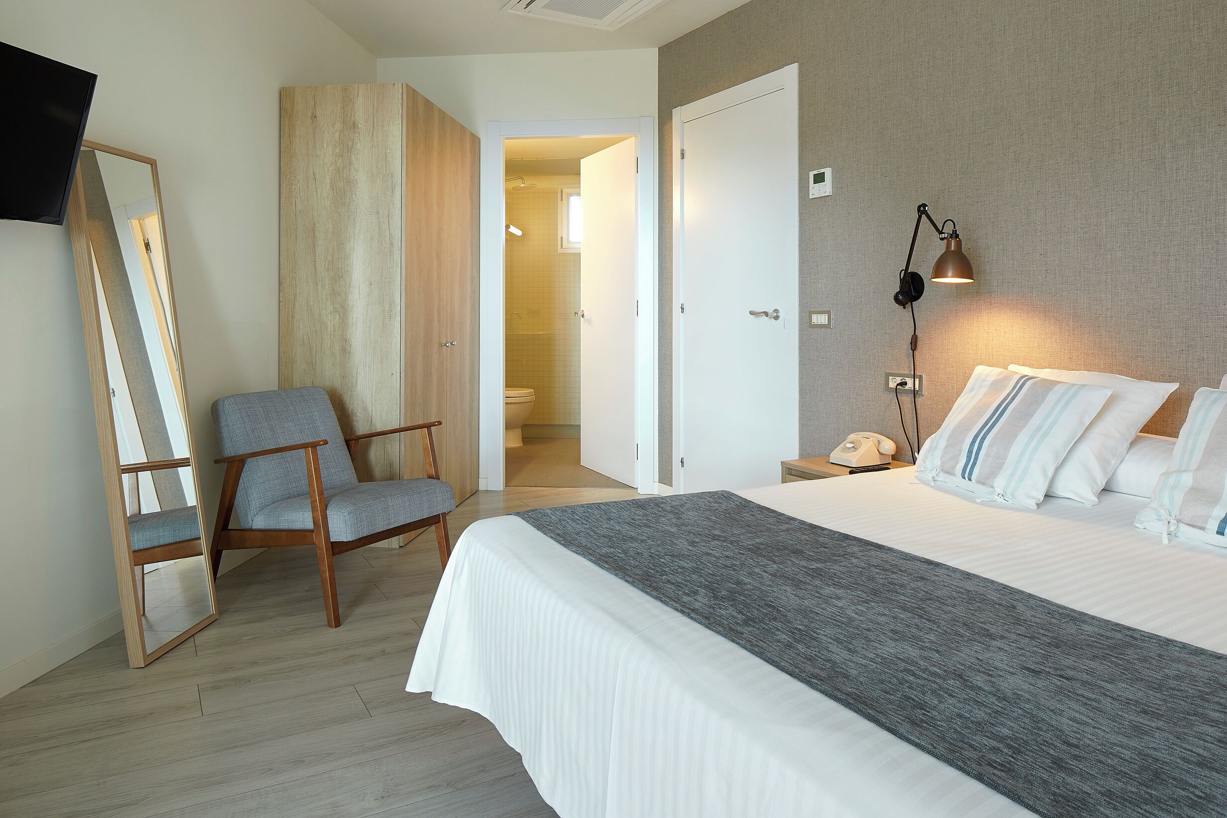 Habitación de hotel con cama, una silla, mesita de noche con lámpara y baño visible a través de una puerta abierta.