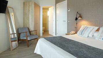 Habitación de hotel con cama, una silla, mesita de noche con lámpara y baño visible a través de una puerta abierta.
