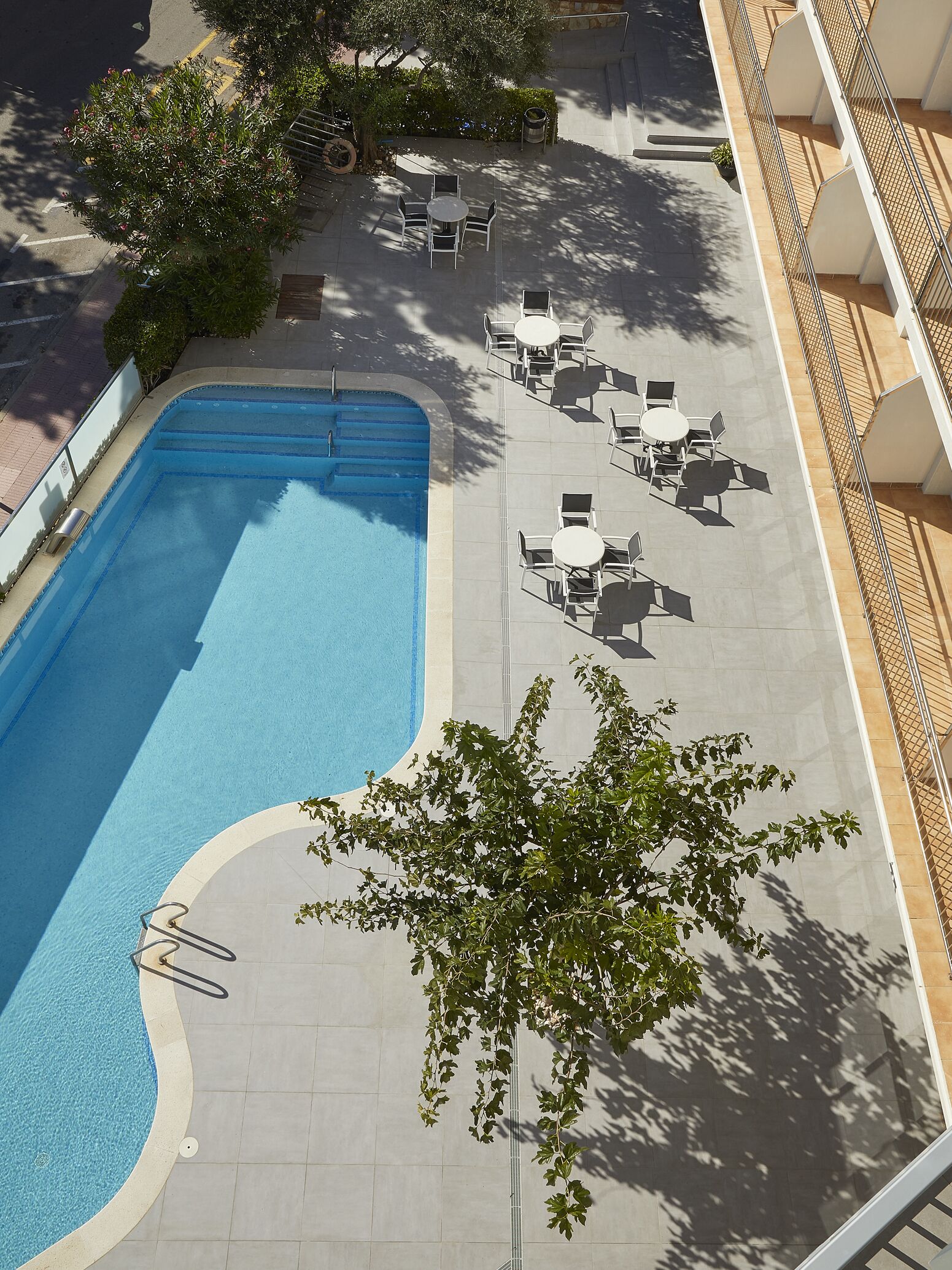 An outdoor swimming pool with adjacent lounge chairs and tables, surrounded by trees and a building with a tiled floor.