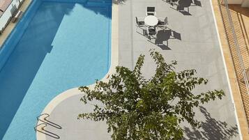 An outdoor swimming pool with adjacent lounge chairs and tables, surrounded by trees and a building with a tiled floor.