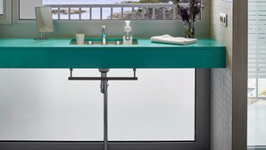 A bathroom with a view of the sea, a teal countertop, a mirror, a sink, and a small vase with flowers.