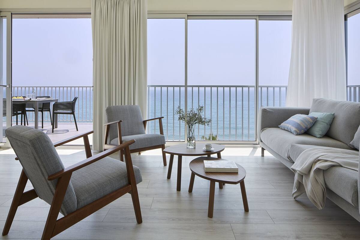 A modern living room with a balcony overlooking the sea, featuring a sofa, chairs, and a small table.