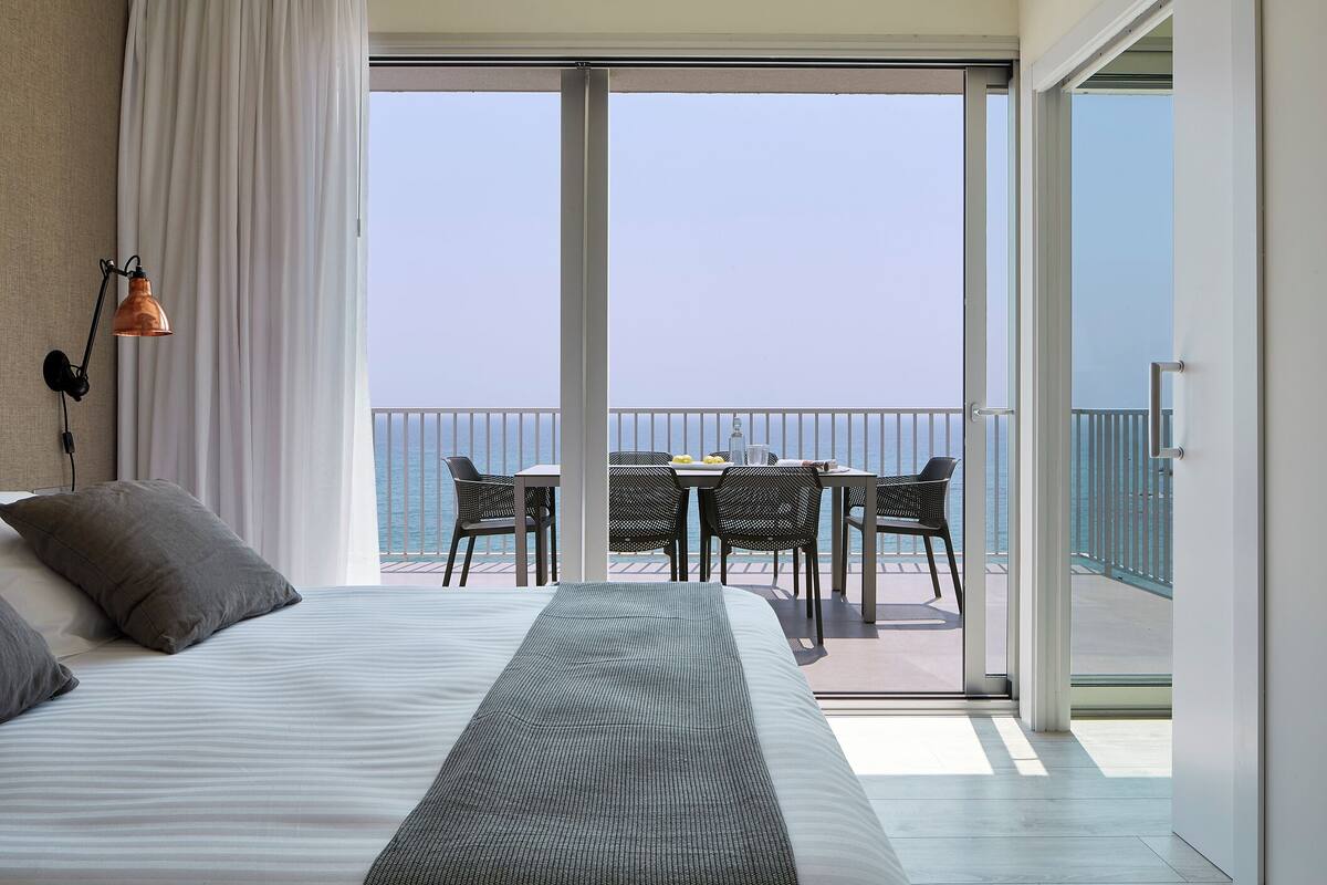 A hotel room with a bed, a table with chairs, and a view of the sea through large glass doors.