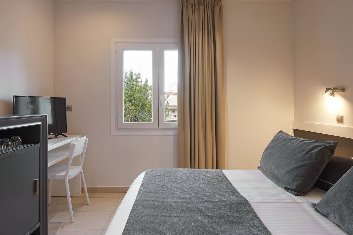 A hotel room with a bed, a desk with a TV, a window with curtains, and a wall-mounted light.