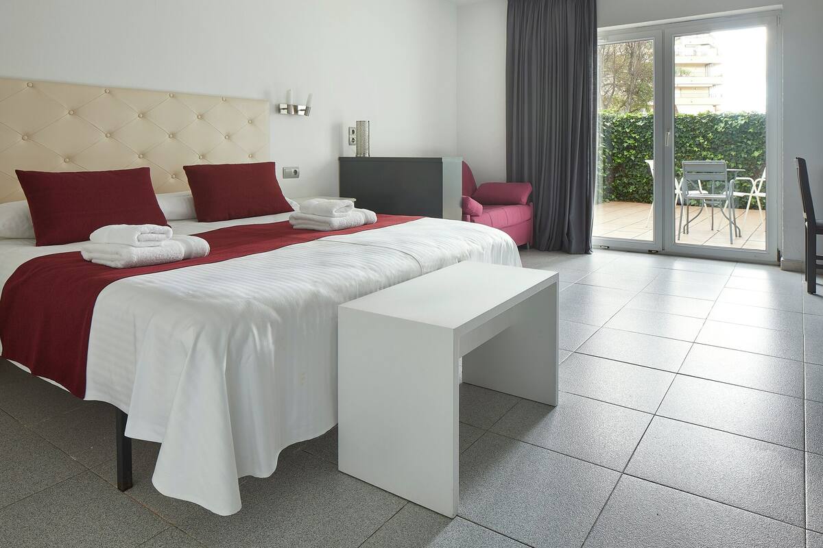 A hotel room with a bed, a desk, a chair, a TV, and a balcony with a table and chairs.