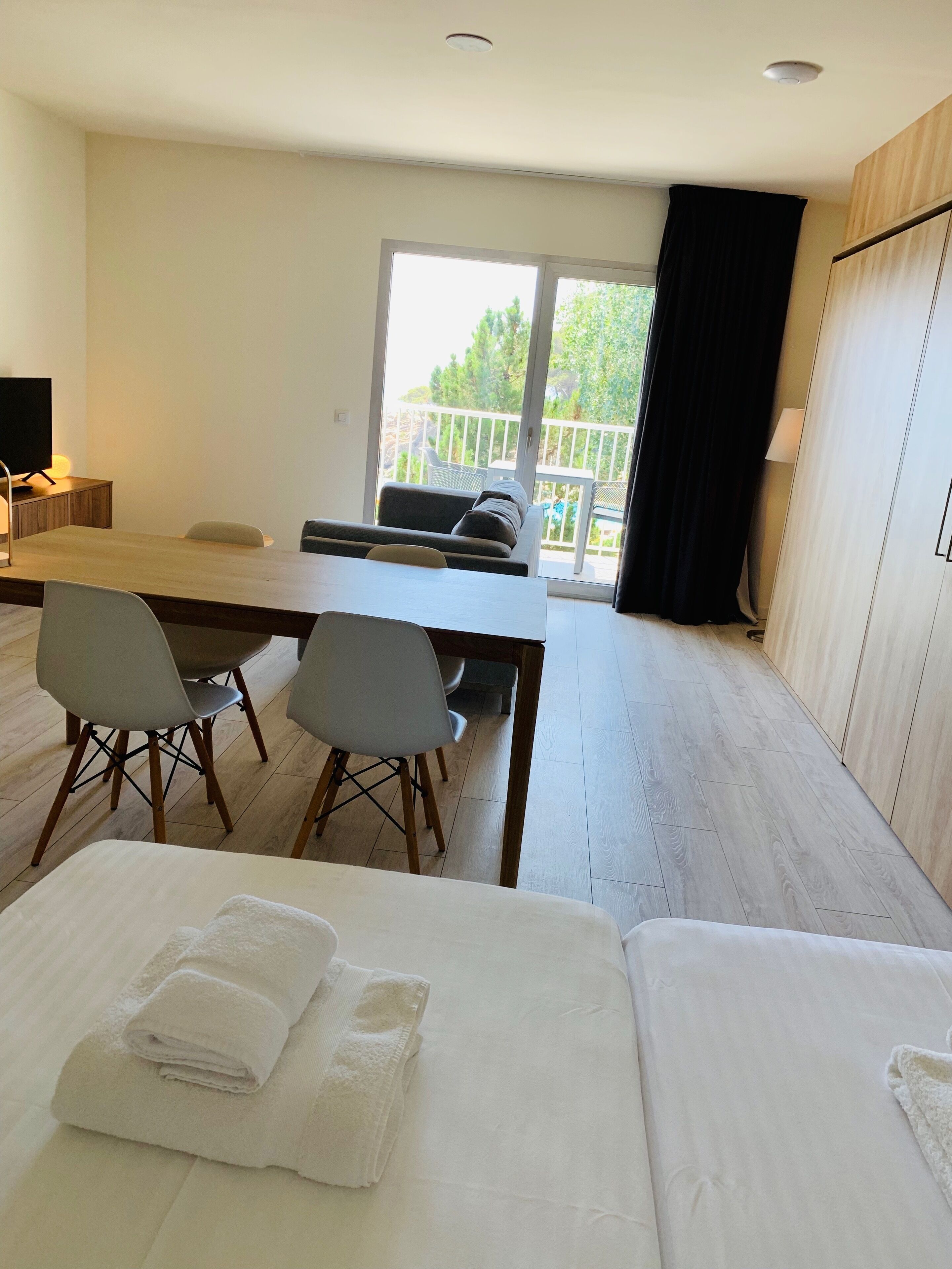 Habitación de hotel con cama, una mesa de comedor con sillas, televisor y balcón con vista a árboles.
