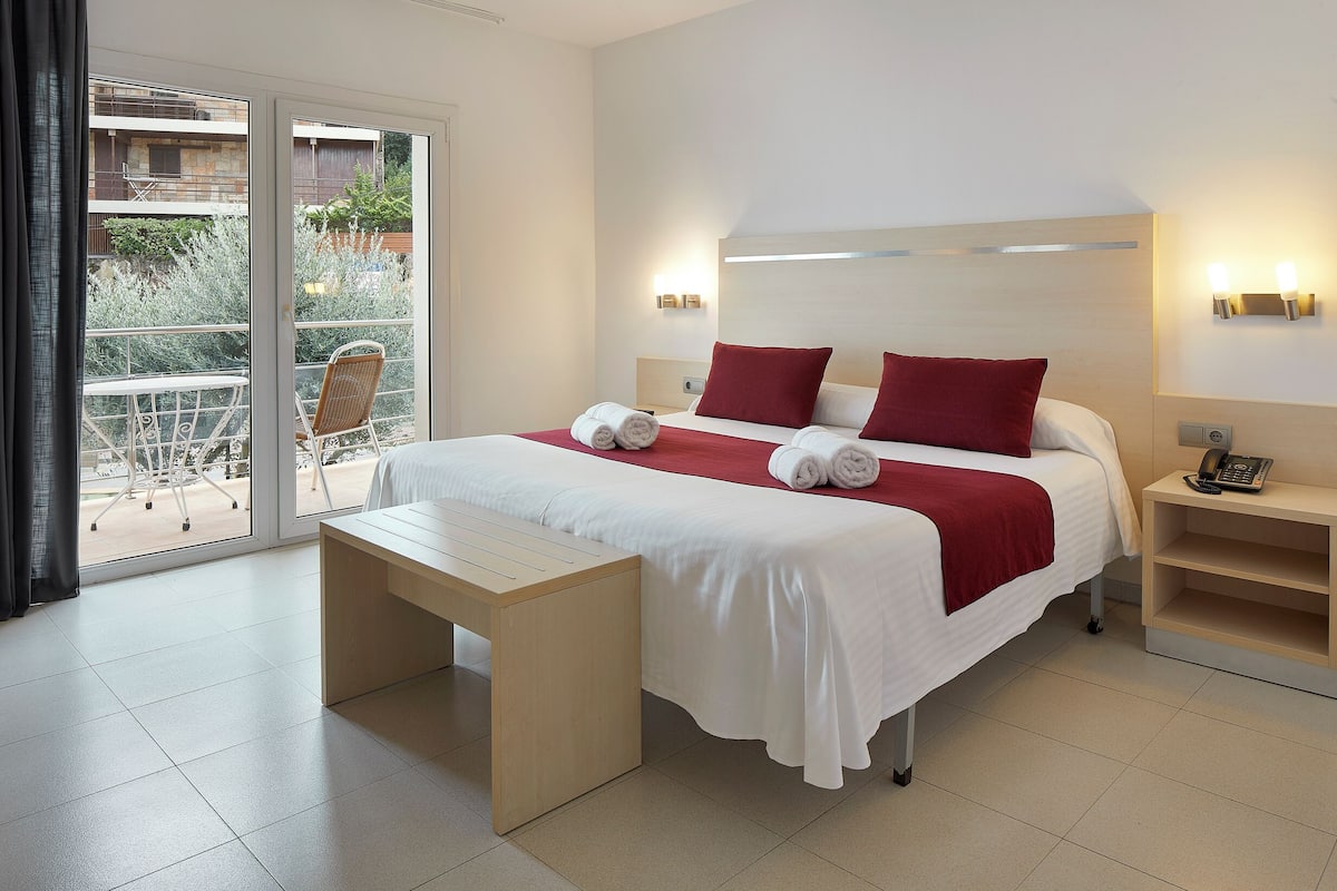 A hotel room with a bed, a bedside table, a nightstand, a bench, and a view of the outside.