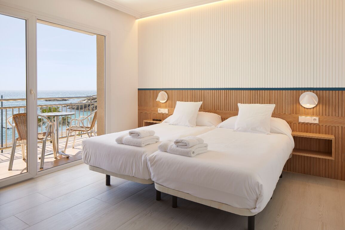 A hotel room with two beds, a balcony with a view of the sea, and a wooden headboard.