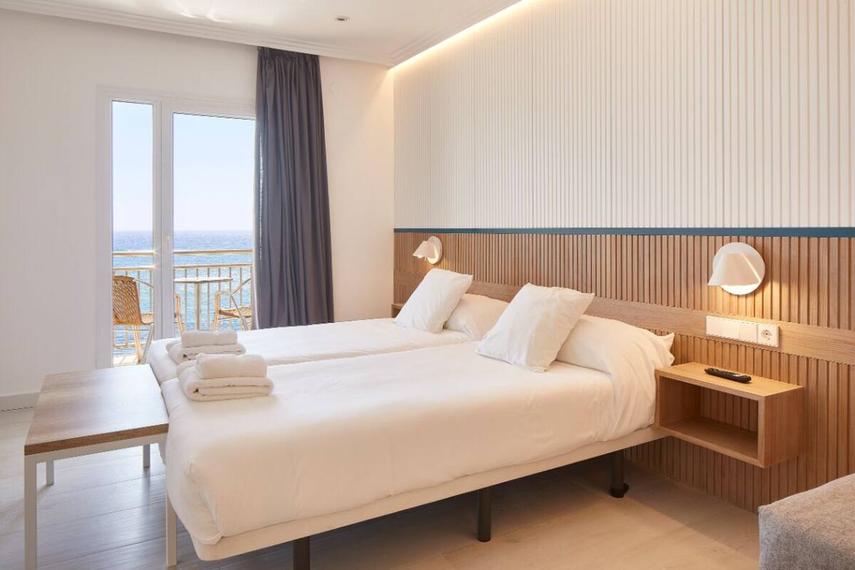 A hotel room with two beds, a wooden headboard, a bedside table with a remote control, and a view of the sea through a large window.