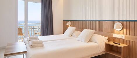 A hotel room with two beds, a wooden headboard, a bedside table with a remote control, and a view of the sea through a large window.