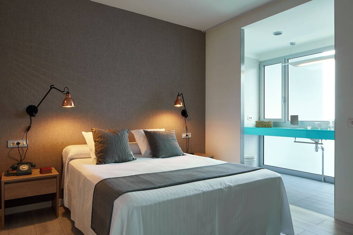 A modern bedroom with a large bed, two bedside lamps, a wooden nightstand, and a view of a balcony through a large window.