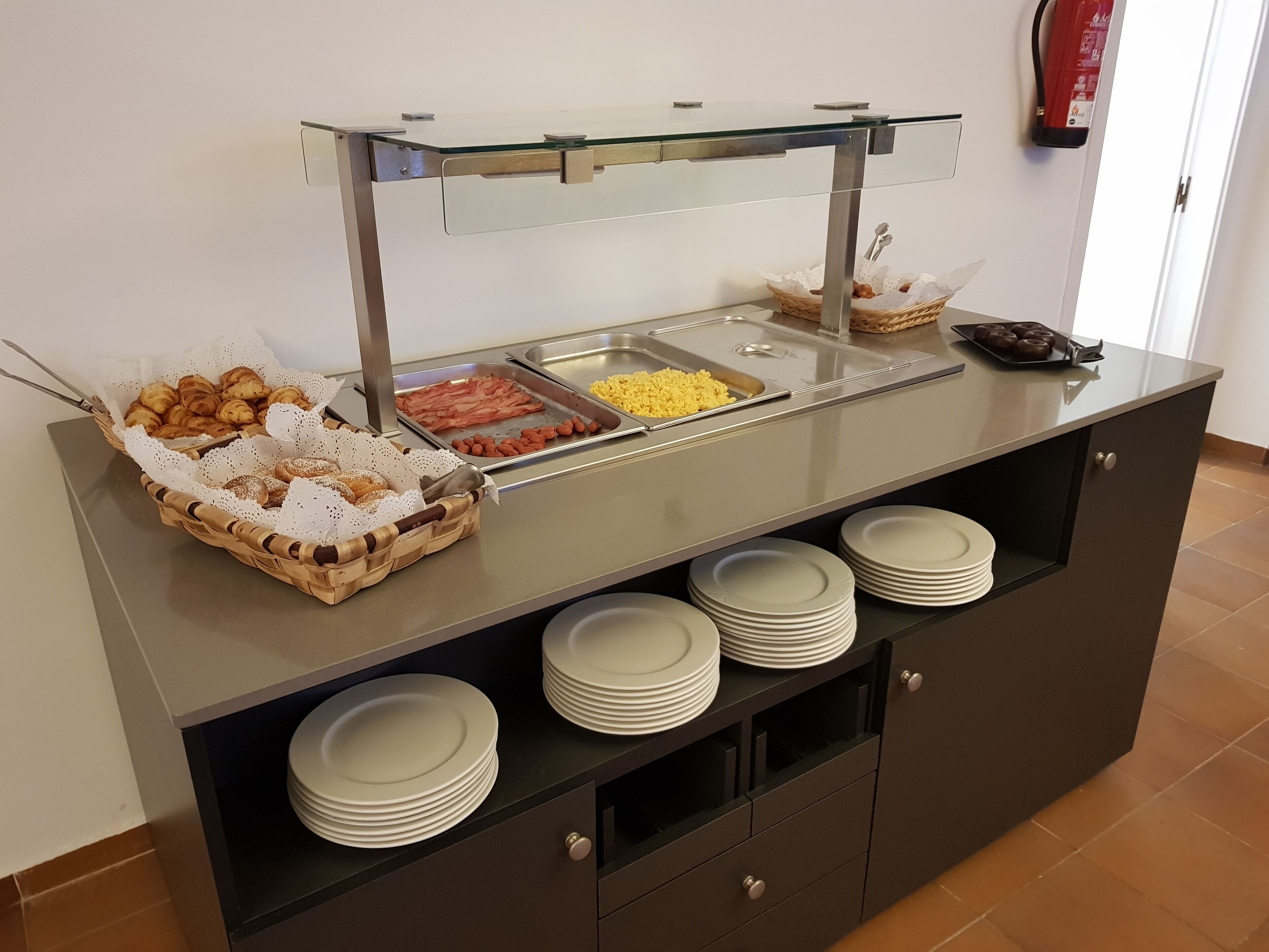 A buffet with various food items, including pastries, bacon, and eggs, and a fire extinguisher mounted on the wall.
