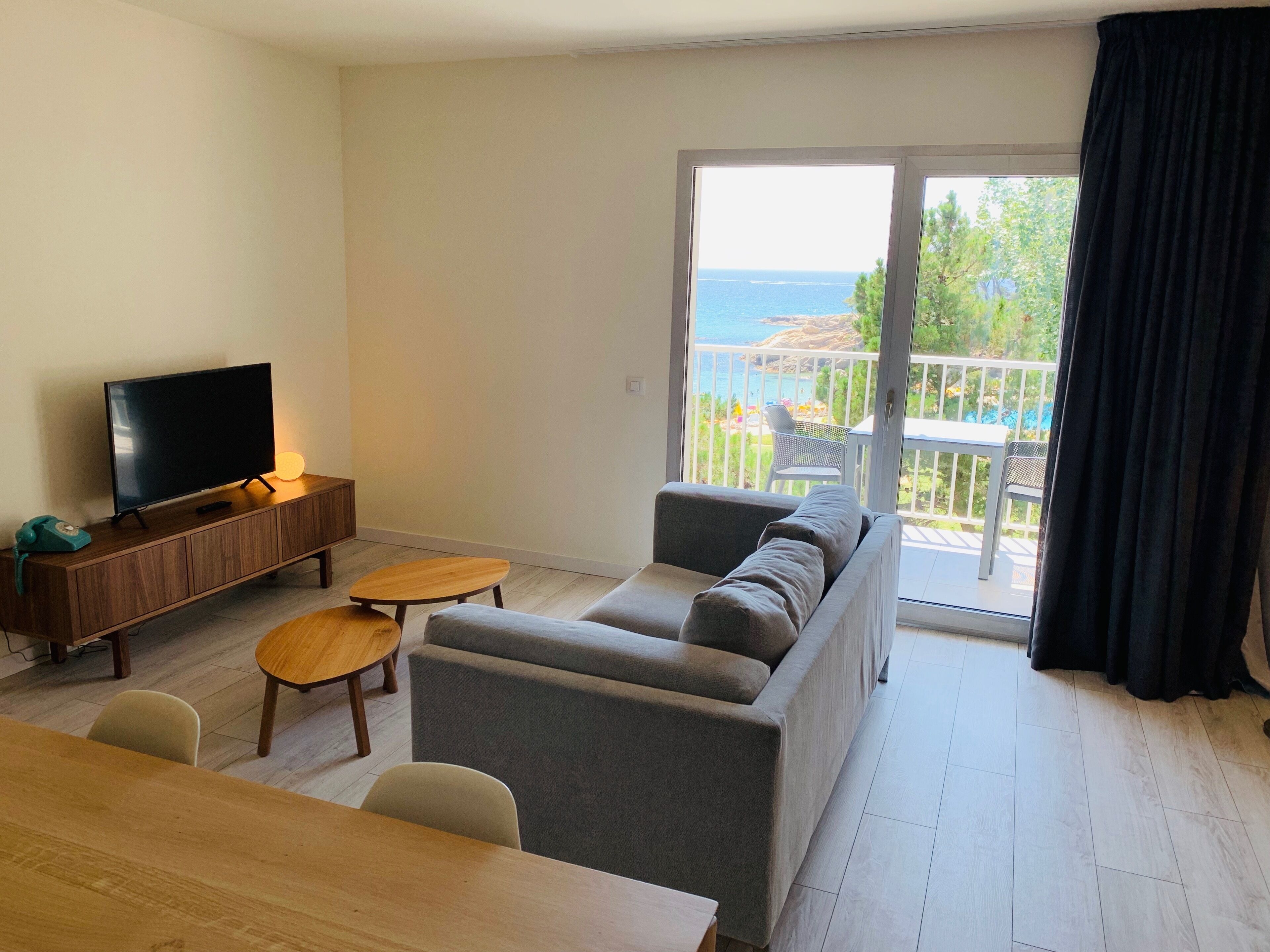 Un moderno salón con un sofá gris, una mesa de centro de madera y un televisor de pantalla plana sobre un mueble de madera. Se ve un balcón con vistas al mar a través de puertas de cristal.