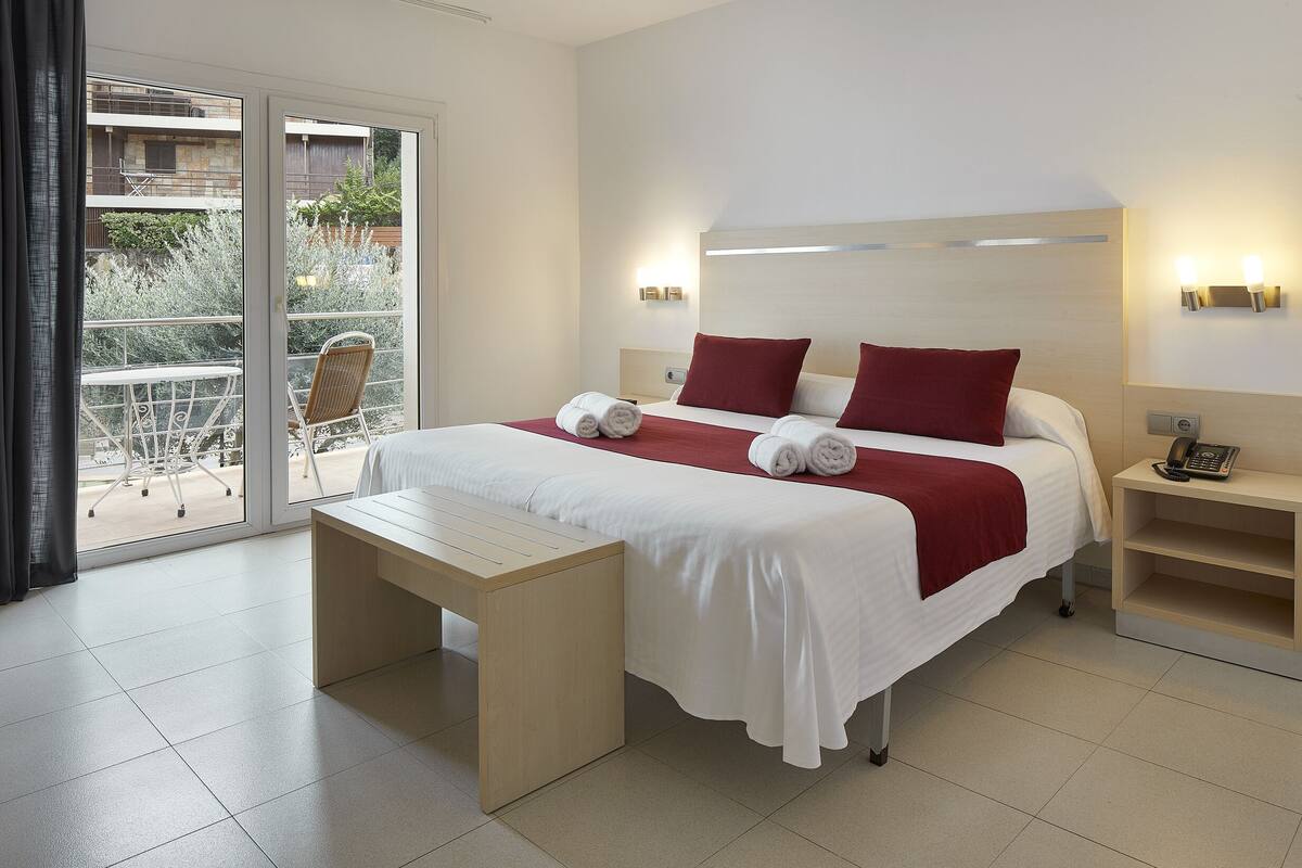 A hotel room with a bed, a bedside table, a desk, and a view of the outside balcony.
