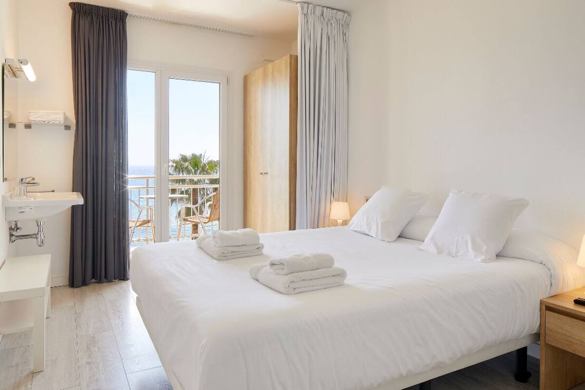 A hotel room with a large bed, a bedside table, a nightstand, a bathroom with a sink, and a view of the sea through a balcony.