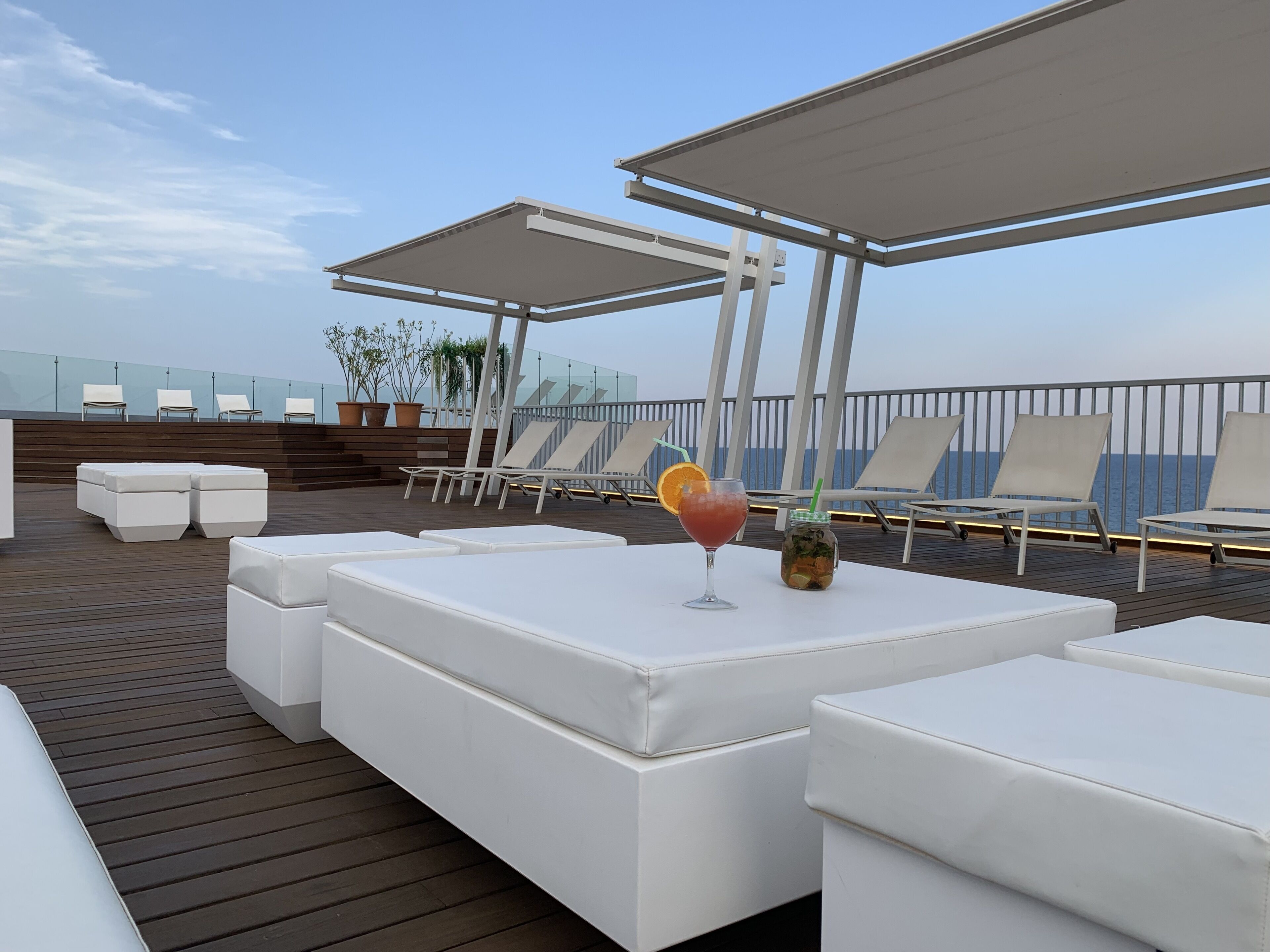 Un espace de détente aménagé sur le toit, avec des sièges blancs et confortables, une boisson servie avec une paille, et une vue sur la mer.