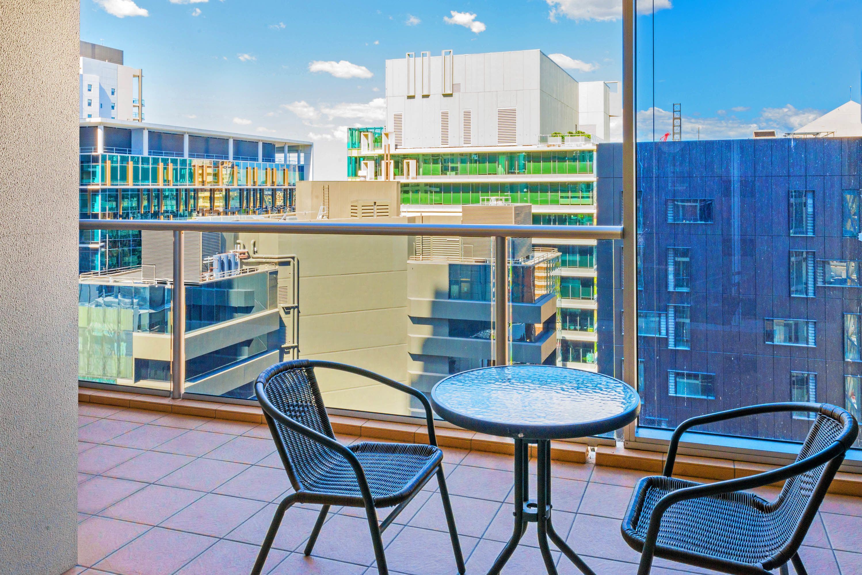 A balcony with two wicker chairs and a round table, overlooking a modern building with multiple floors and large windows.