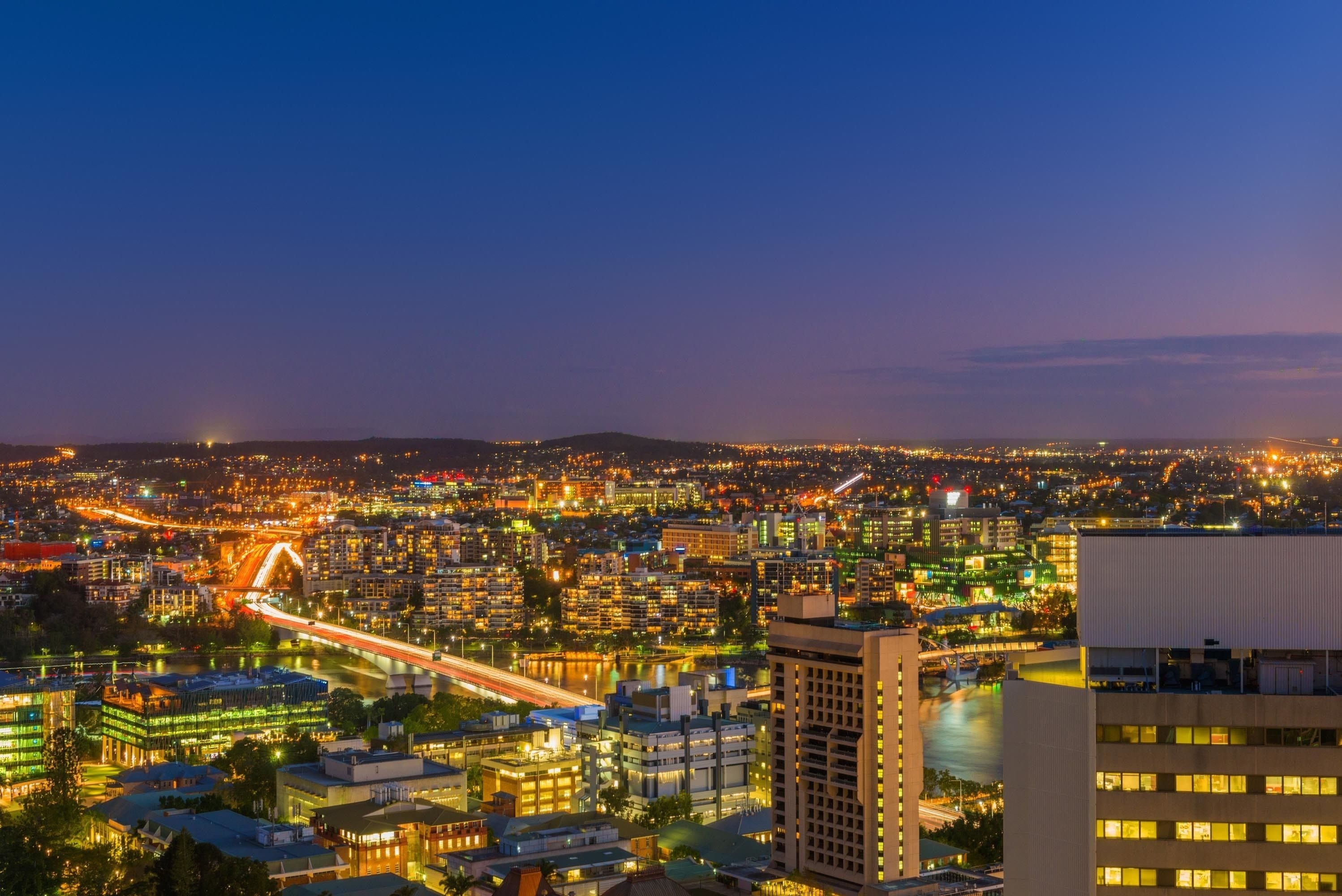 A cityscape at dusk with illuminated buildings, a river, and a bridge.
