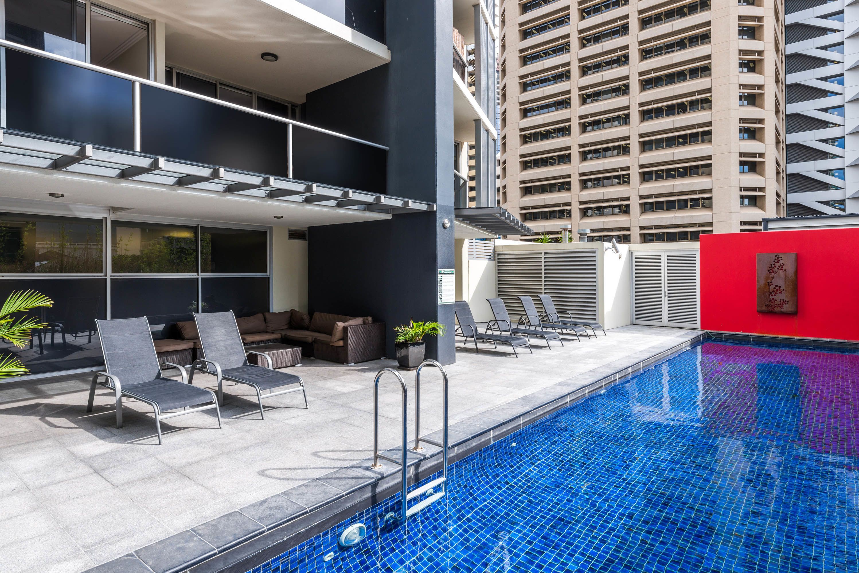 A rooftop pool area with lounge chairs, a small seating area, and a view of a tall building in the background.