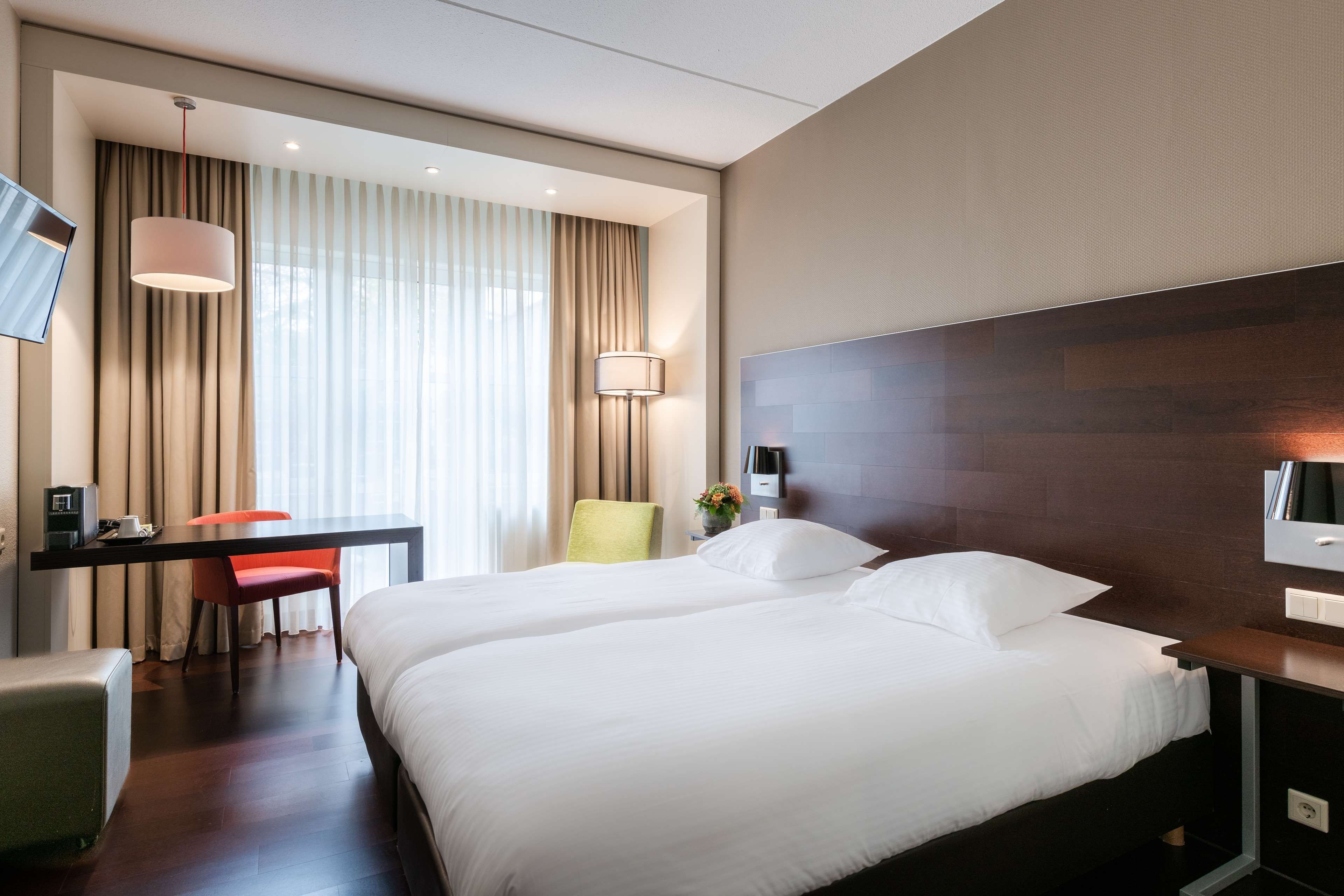 Habitación de hotel con cama, mesitas de noche, un escritorio con silla, lámpara y televisor montado en la pared.