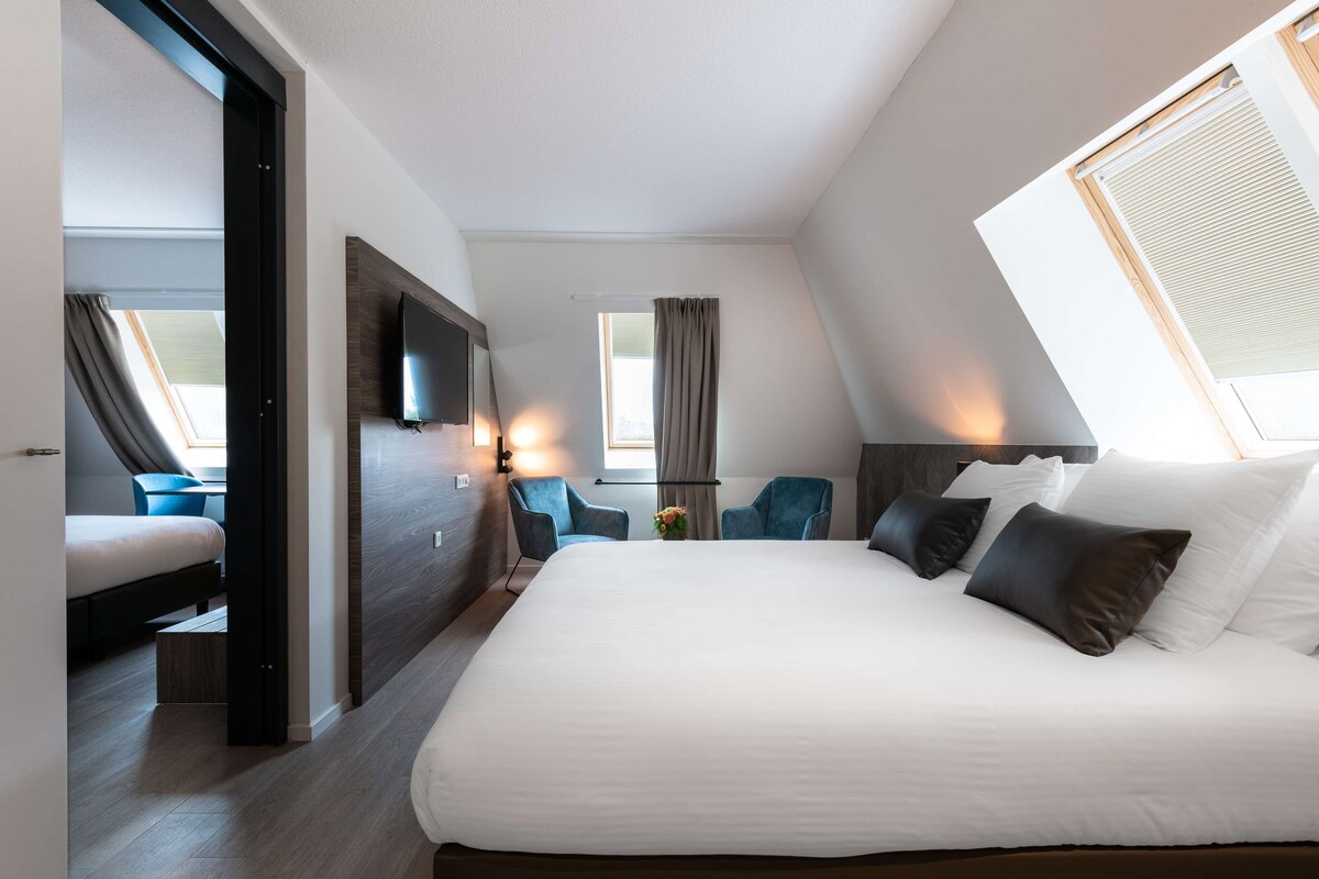 A modern hotel room with a large bed, a flat-screen TV, and a view of the city through a window.