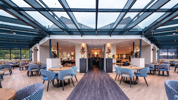 Un vaste espace de restauration intérieur, doté d’un haut plafond et de panneaux de verre, aménagé avec un mobilier moderne et comprenant une allée centrale.