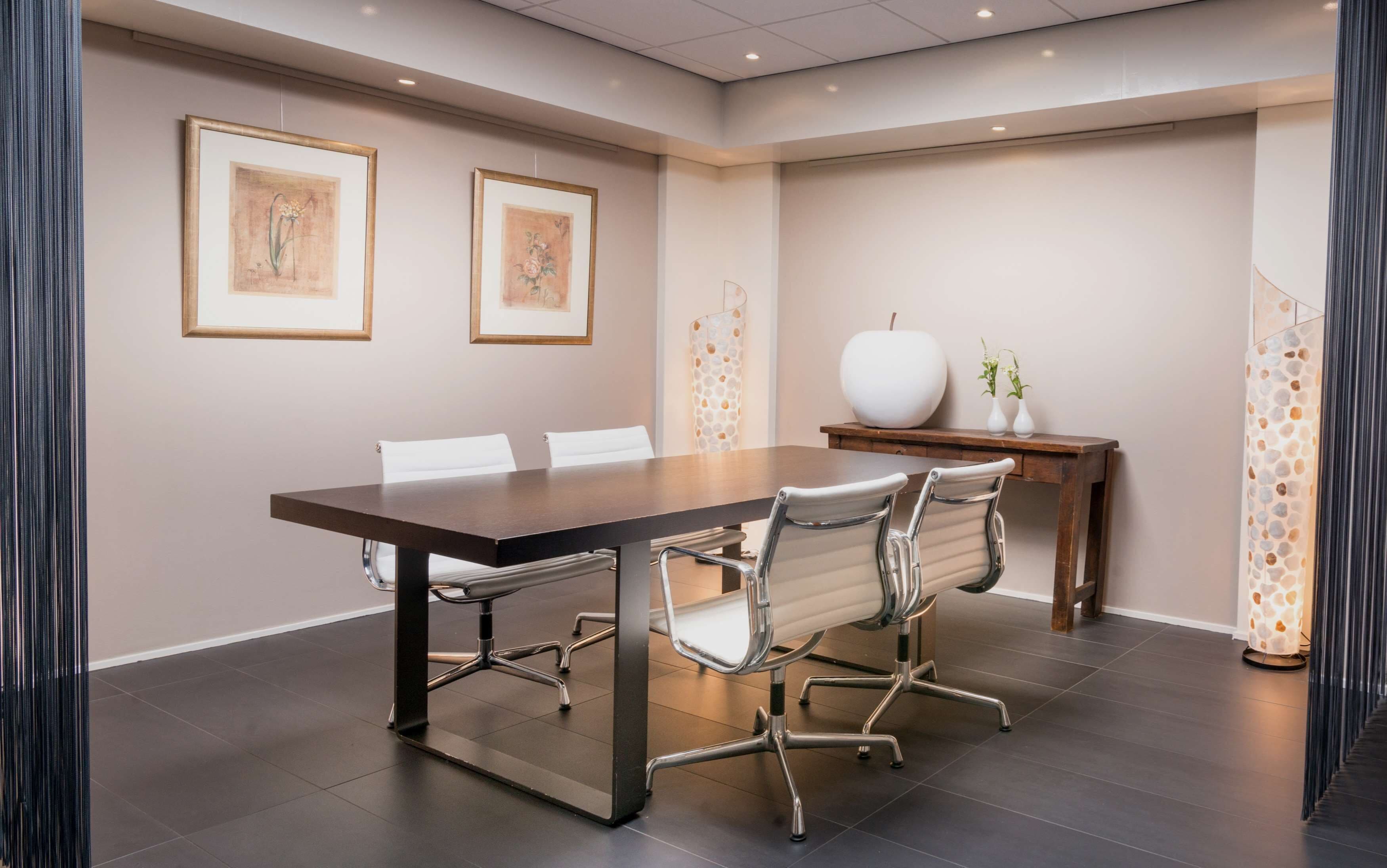 A conference room with a dark wooden table, white chairs, framed floral artwork on the walls, a small white vase, and a decorative light fixture.