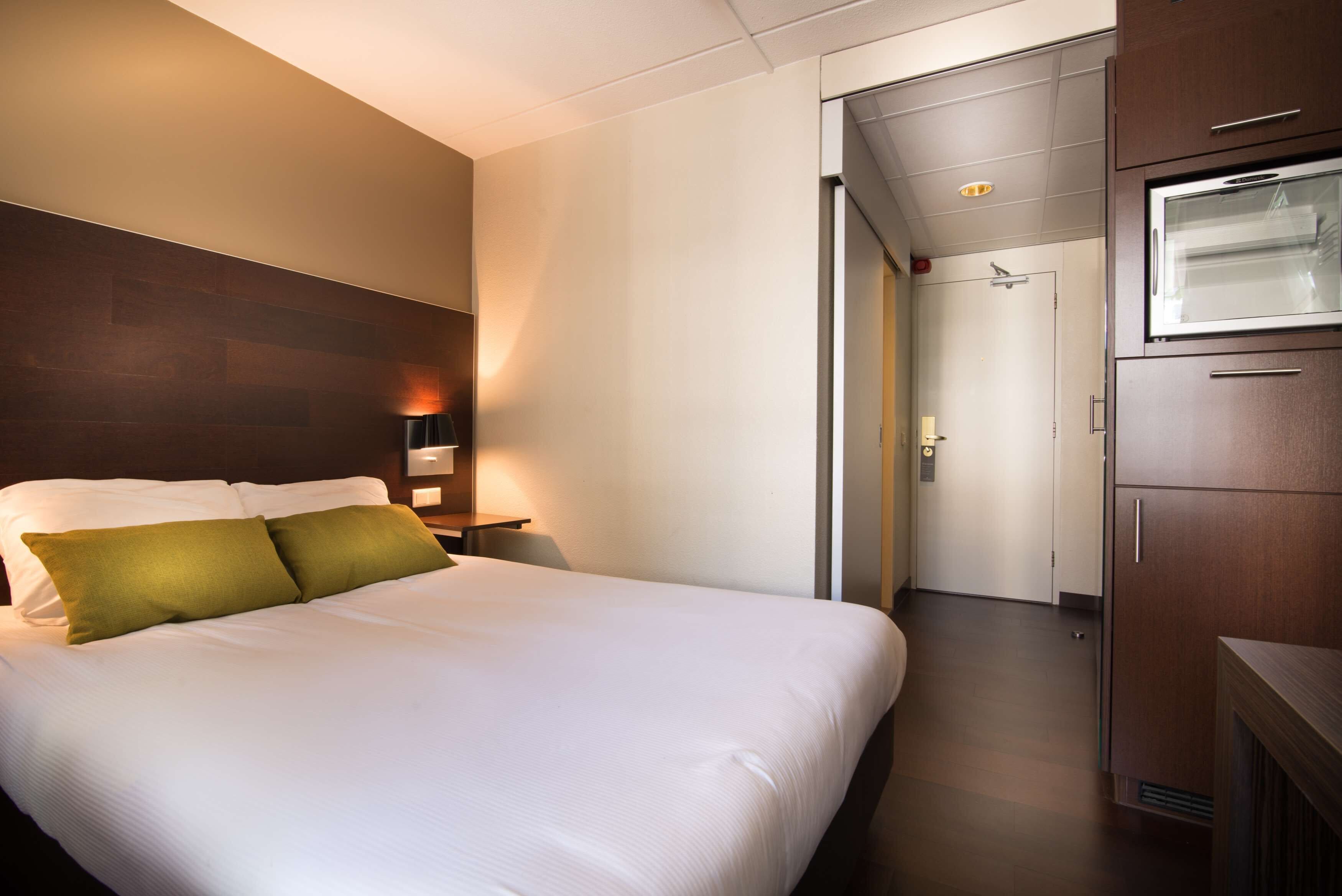 A hotel room with a large bed, a nightstand with a lamp, a wardrobe, and a door leading to a bathroom.