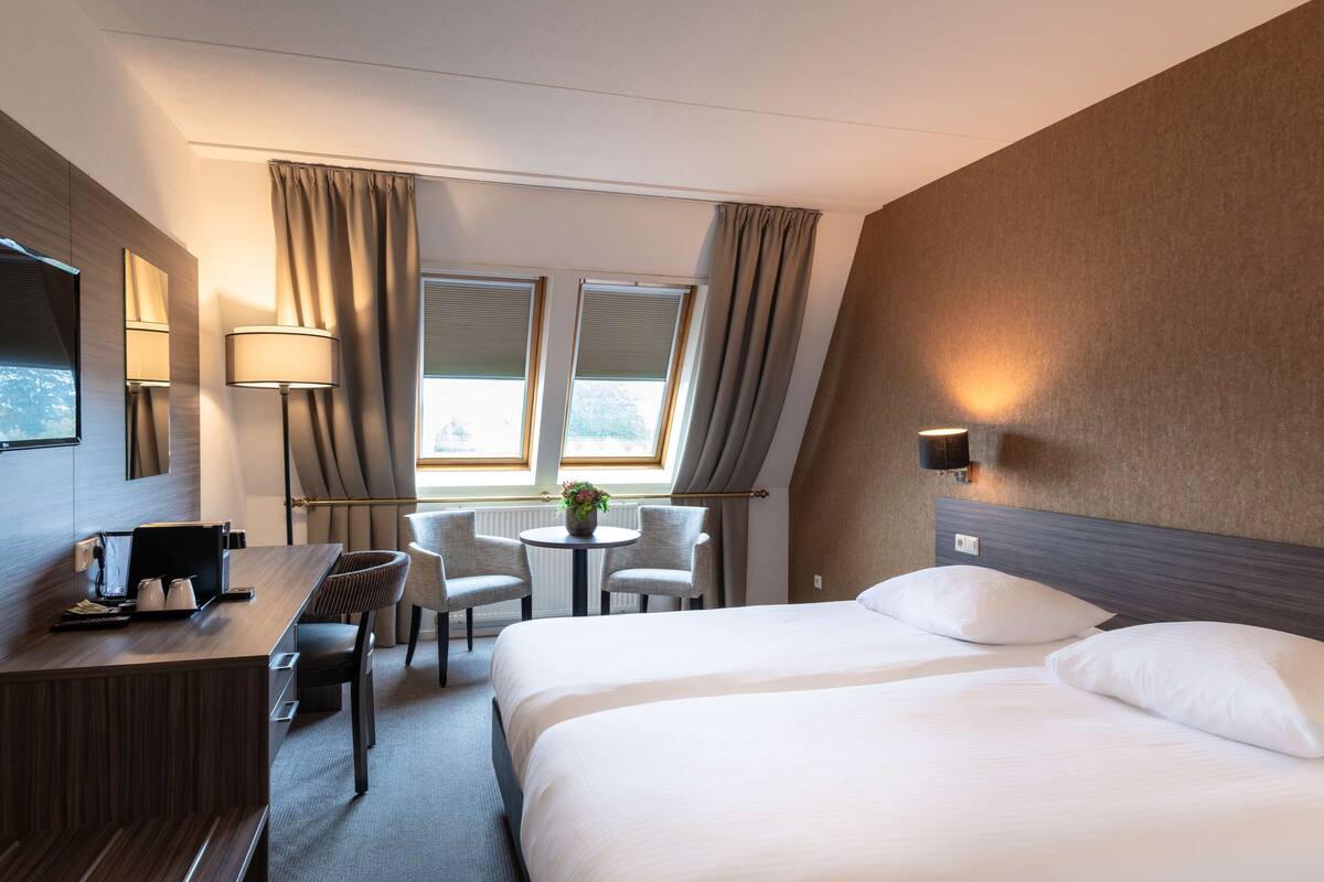 Hotelkamer met een bed, bureau, twee stoelen, een kleintje tafeltje met een plantje en een raam met gordijnen.