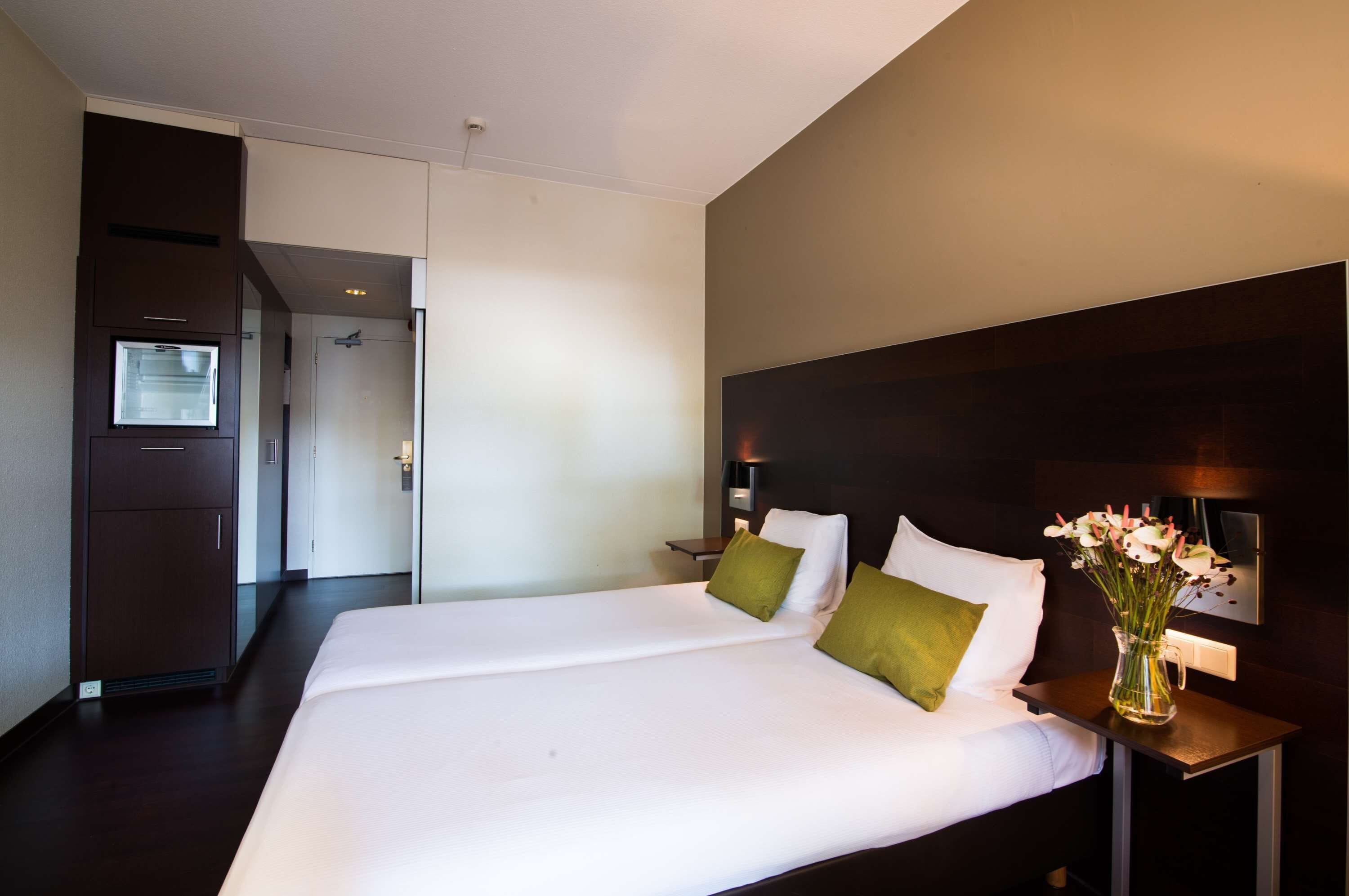 Una habitación de hotel con una cama grande, una mesita de noche, un jarrón con flores y un televisor montado en la pared.