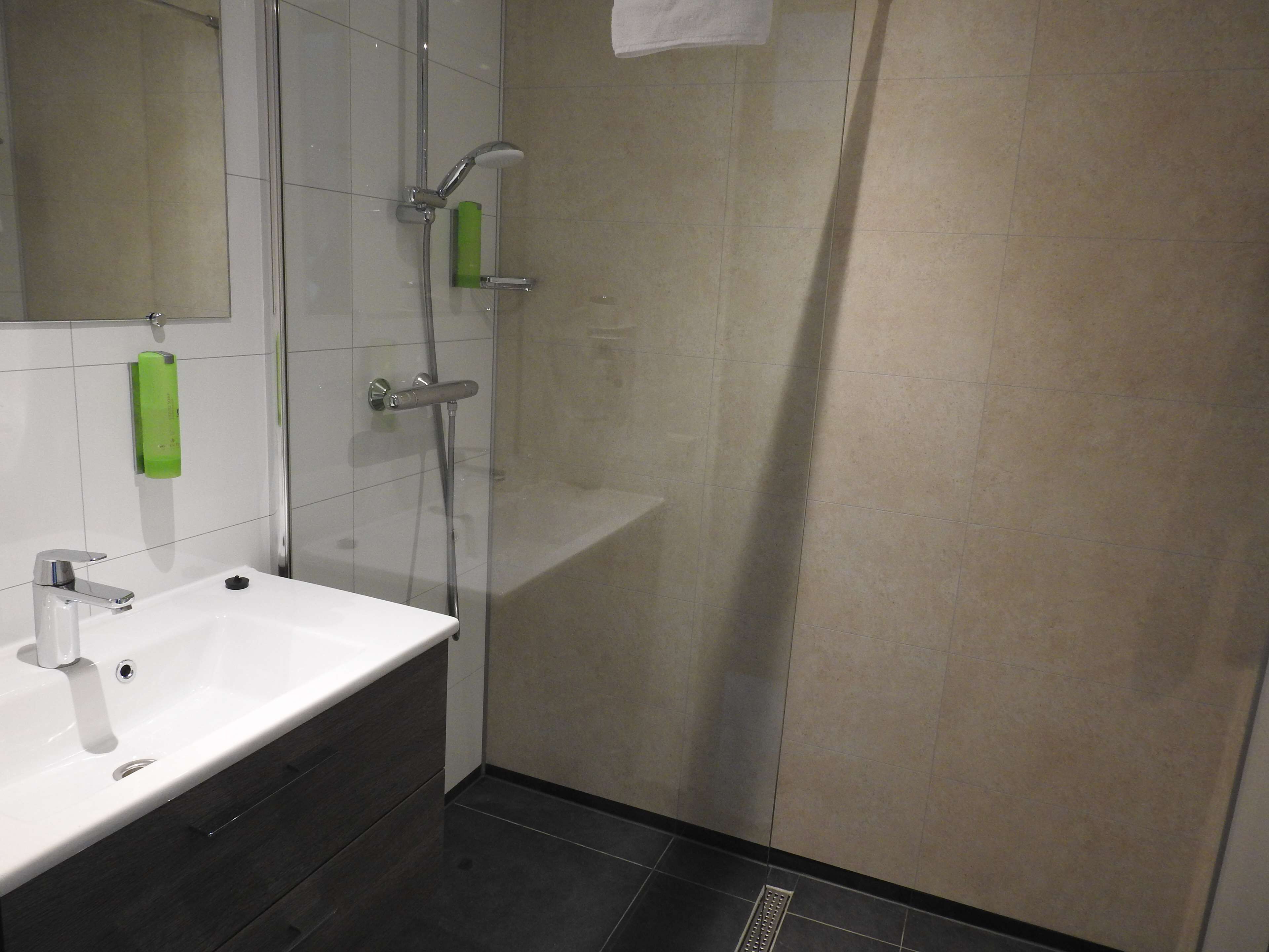 A modern bathroom with a white sink, a mirror, a glass shower enclosure, and a green soap dispenser.