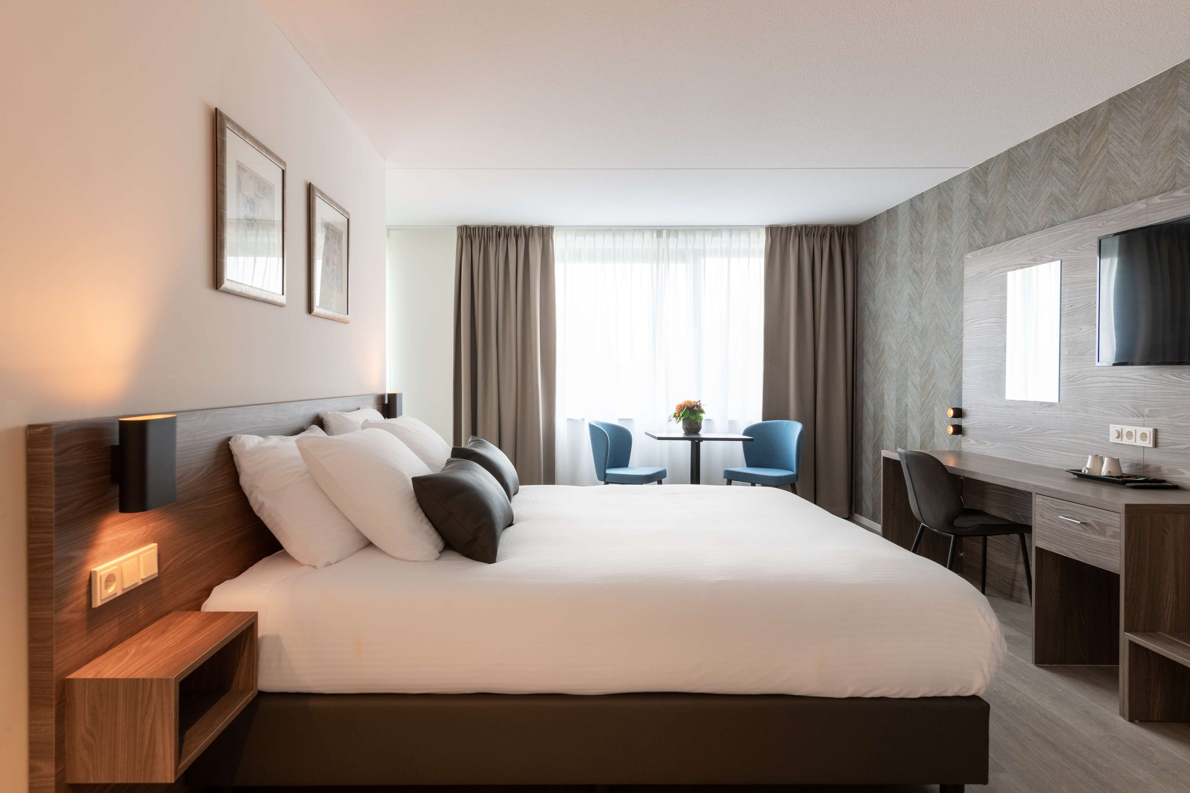 Une chambre d’hôtel avec un grand lit, un bureau, deux chaises, une télévision et une fenêtre avec des rideaux.