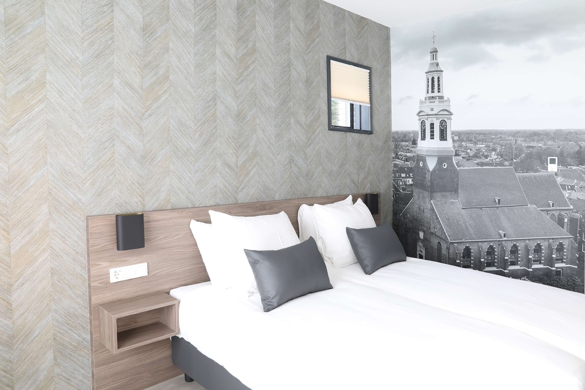 Een moderne hotelkamer met een groot bed, een nachtkastje en uitzicht op een historisch gebouw met een toren.