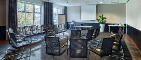 A conference room with rows of chairs, a projector screen, a whiteboard, and a table with a plant.