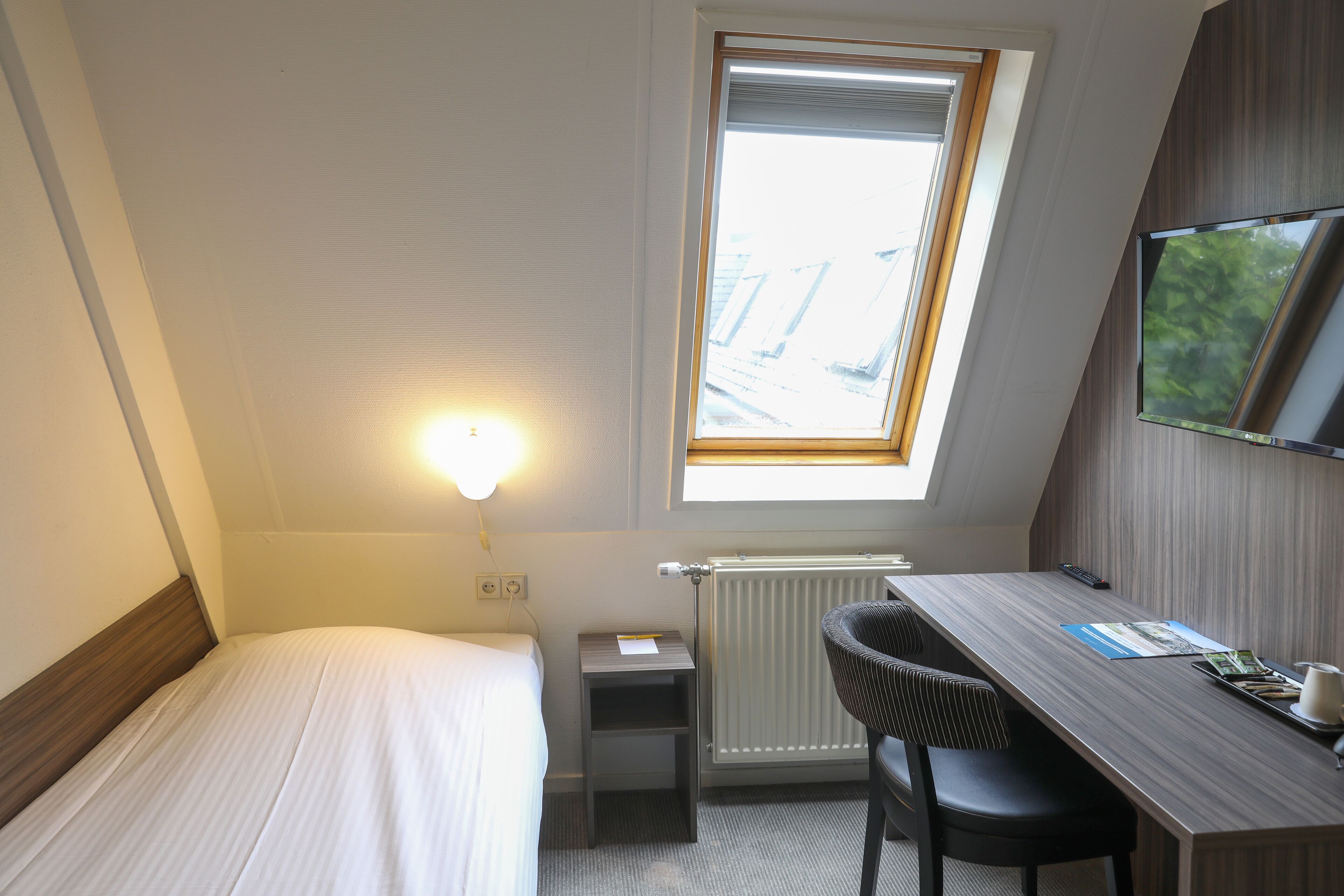 Ein kleines Hotelzimmer mit einem Einzelbett, einem Schreibtisch mit Stuhl, einem an der Wand montierten Fernseher und einem Fenster mit Jalousien.