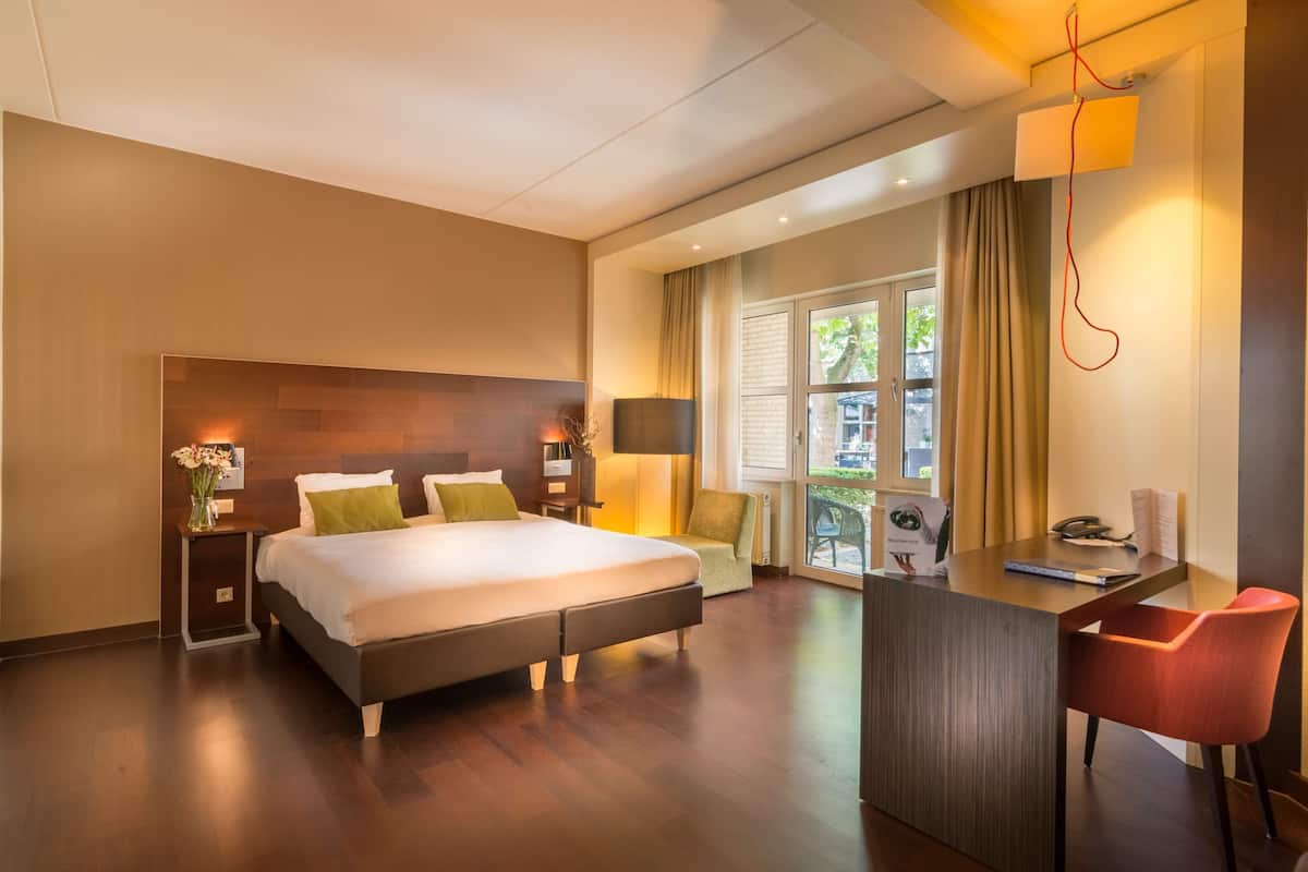 Een moderne hotelkamer met een groot bed, een bureau met stoel, een klein tafeltje met een bloevas en een raam met gordijnen.