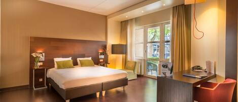 A modern hotel room with a large bed, a desk with a chair, a small table with a vase of flowers, and a window with curtains.