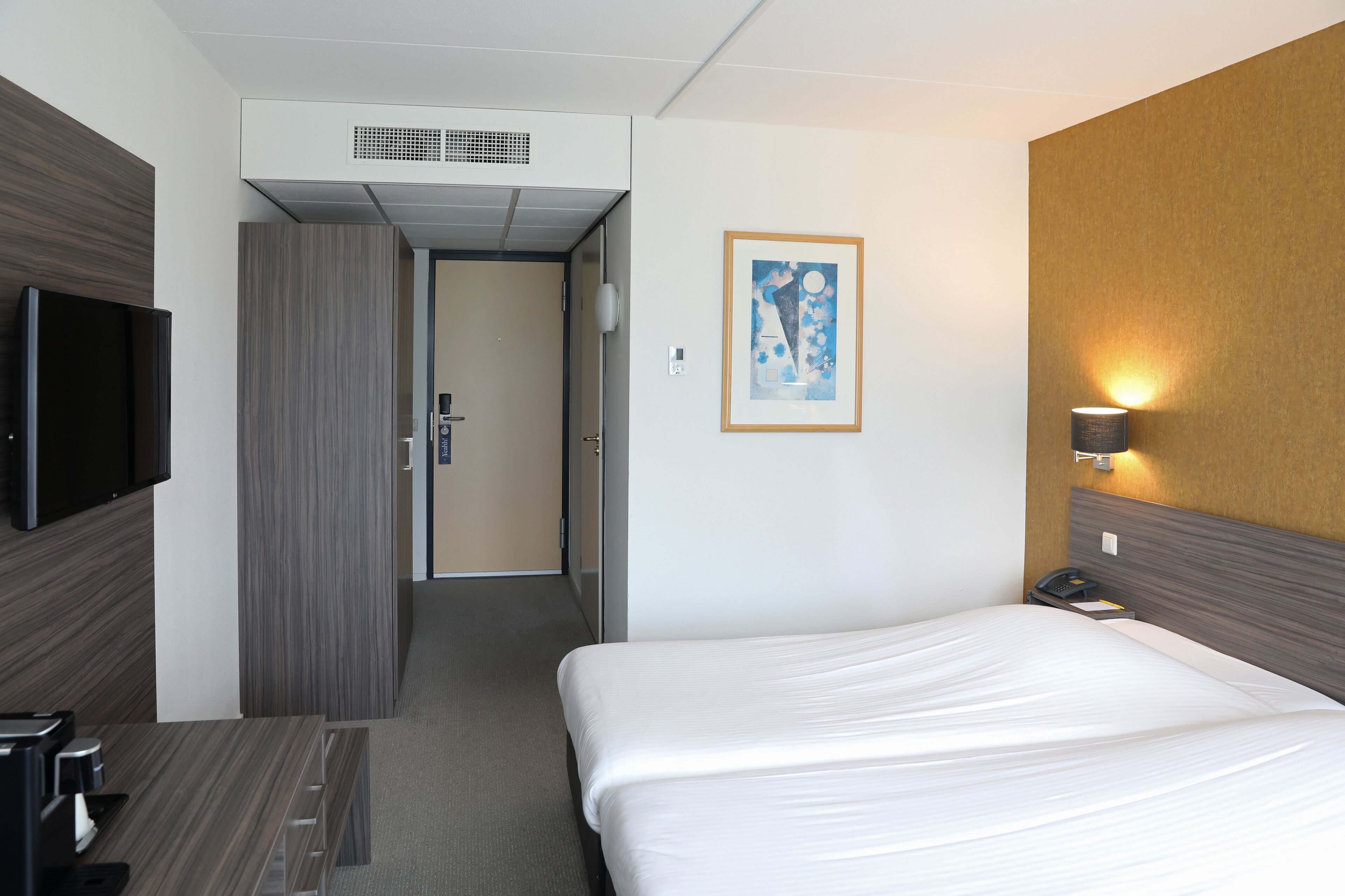 Habitación de hotel con cama, escritorio con cafetera, televisión de pantalla plana, un cuadro enmarcado en la pared y lámpara de pared.