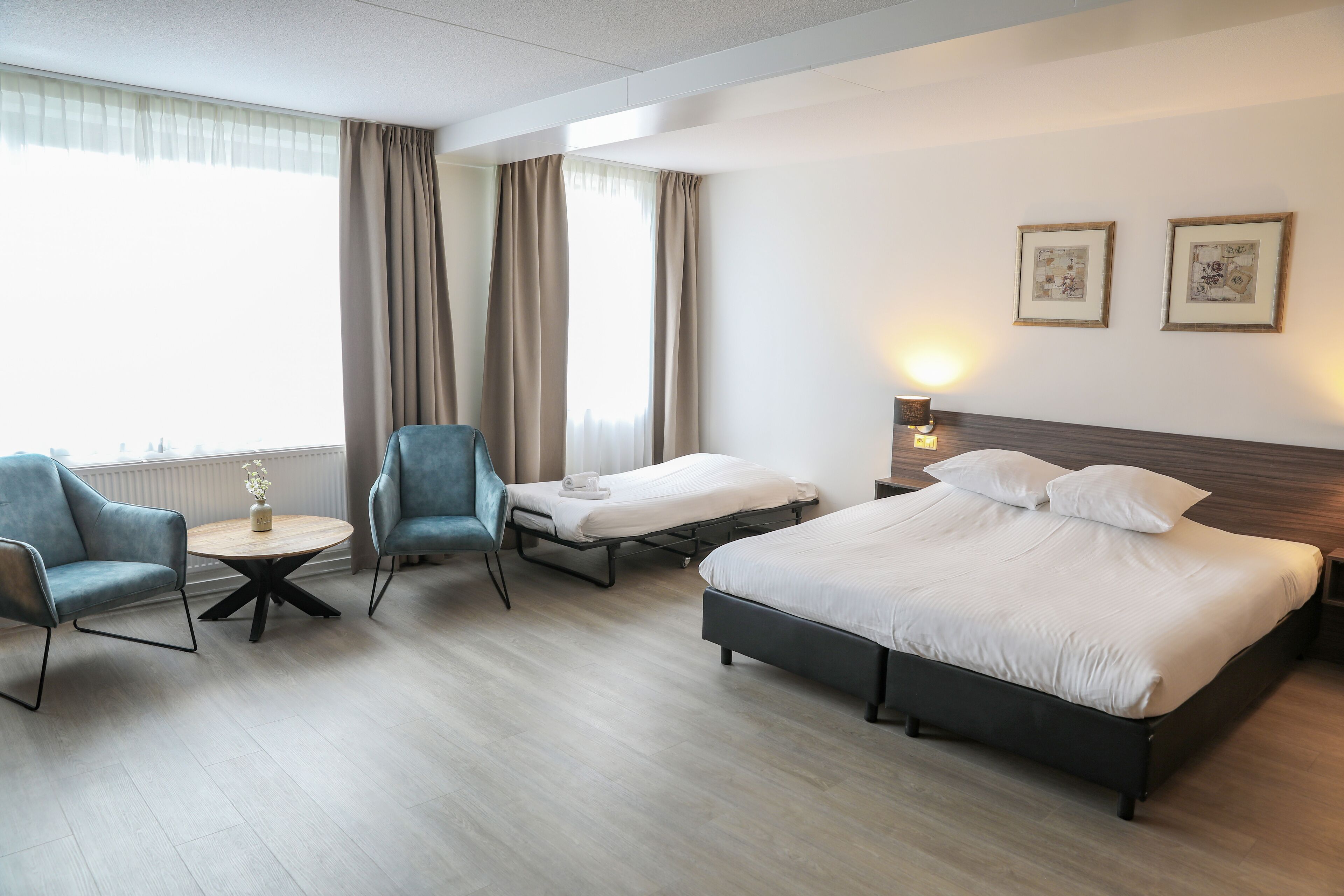 A hotel room with a large bed, two single beds, a round table with a vase, two armchairs, and a window with curtains.