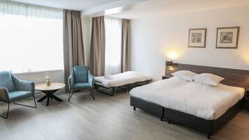 A hotel room with a large bed, two single beds, a round table with a vase, two armchairs, and a window with curtains.