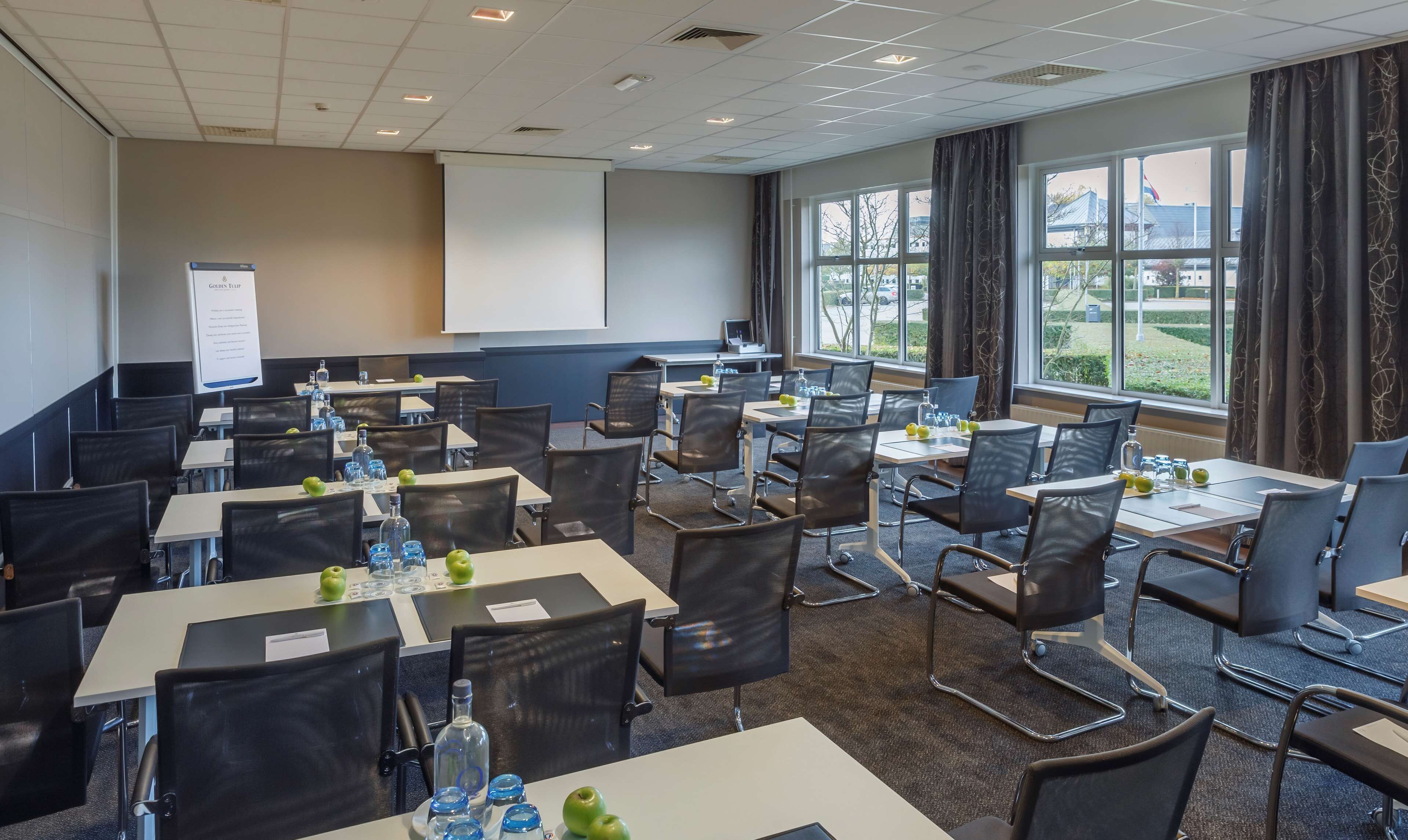 A conference room with tables and chairs, a projector screen, and large windows with curtains.