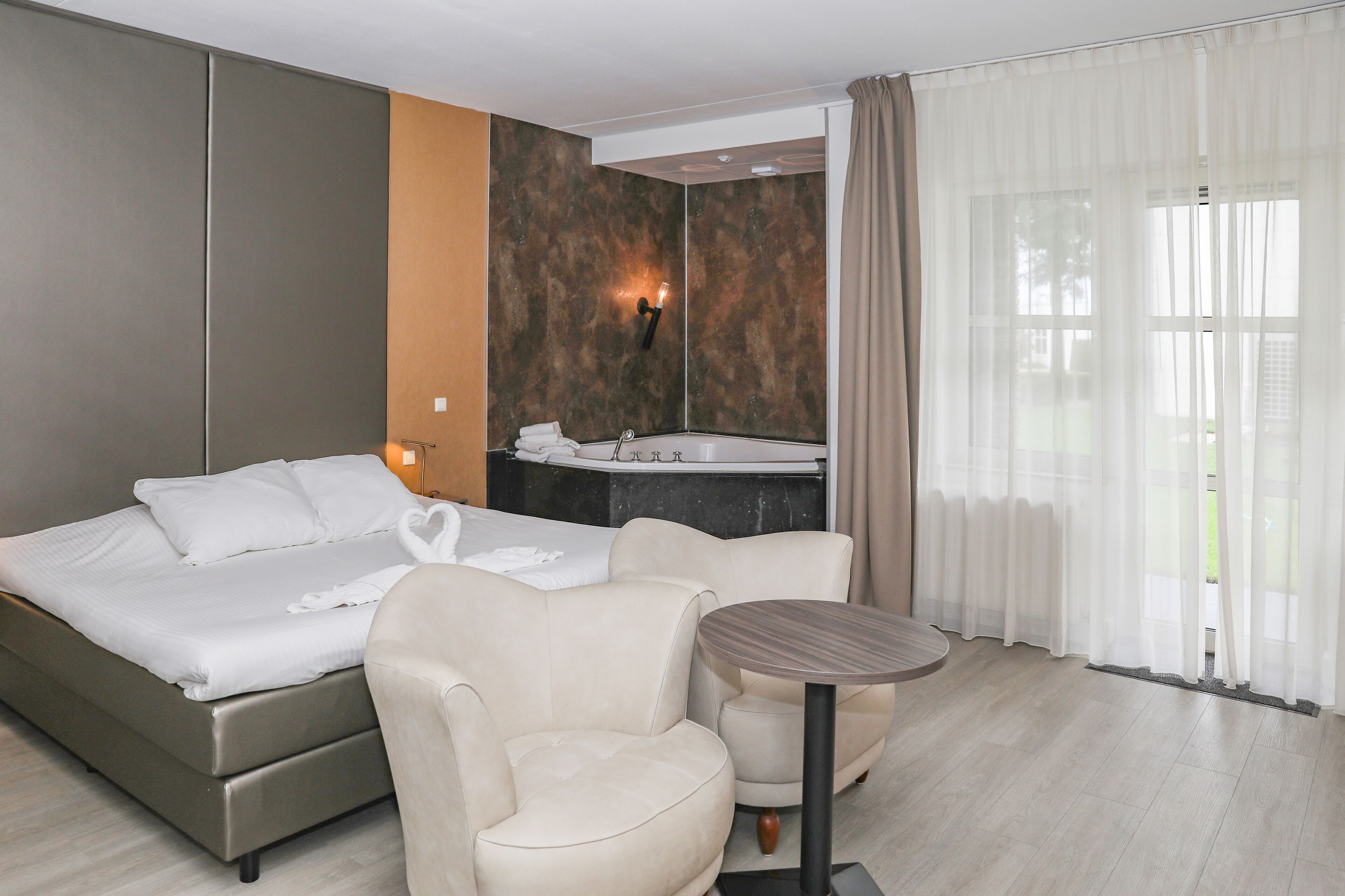A hotel room with a bed, a seating area, a small table, a bathroom with a sink, and a large window with curtains.