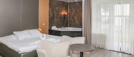 A hotel room with a bed, a seating area, a small table, a bathroom with a sink, and a large window with curtains.
