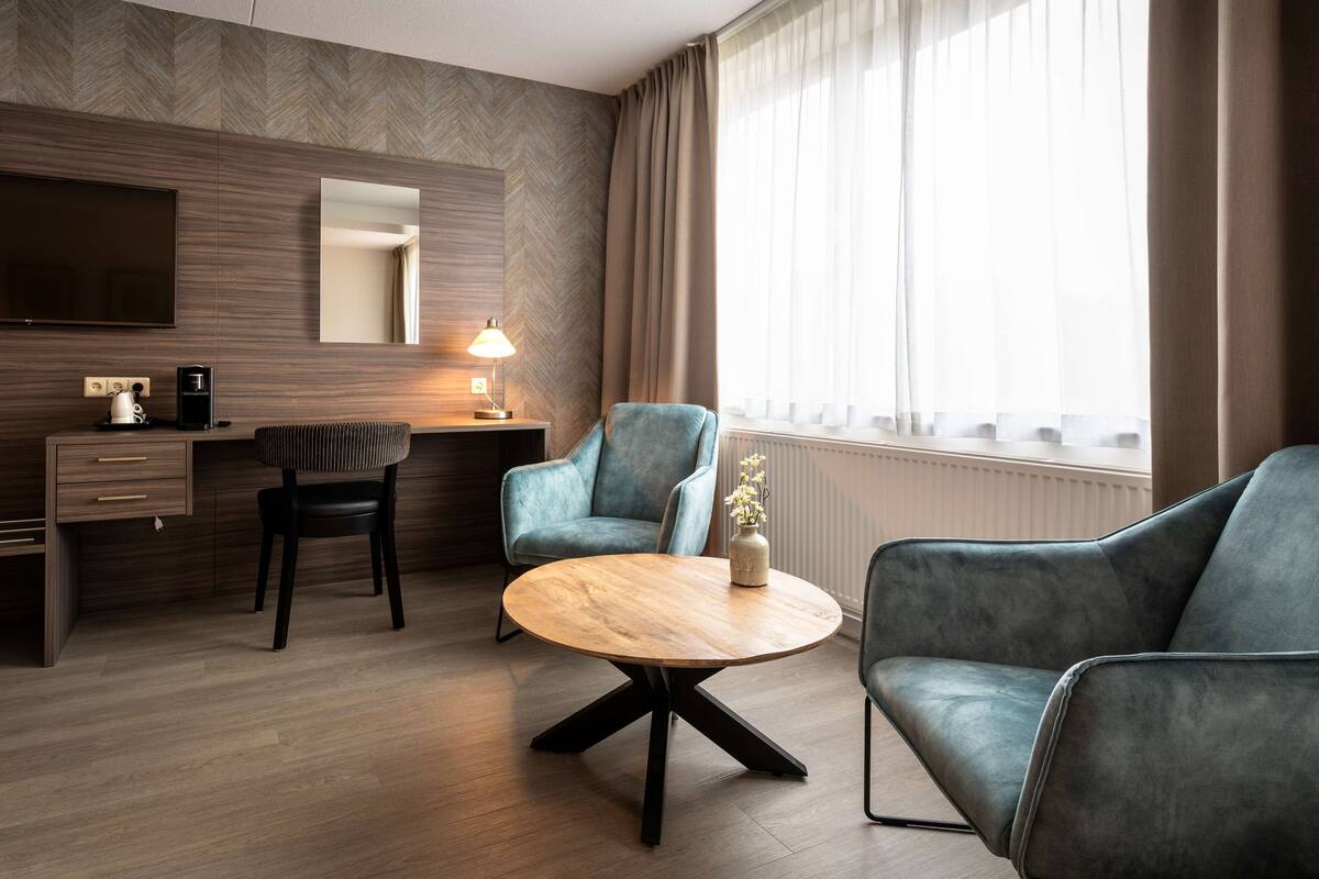 Een moderne hotelkamer met een wand van hout in visgraatmotief, een flatscreen televisie, een bureau met koffiezetapparaat, een ronde houten tafel, twee gestoffeerde fauteuils en een groot raam met gordijnen.