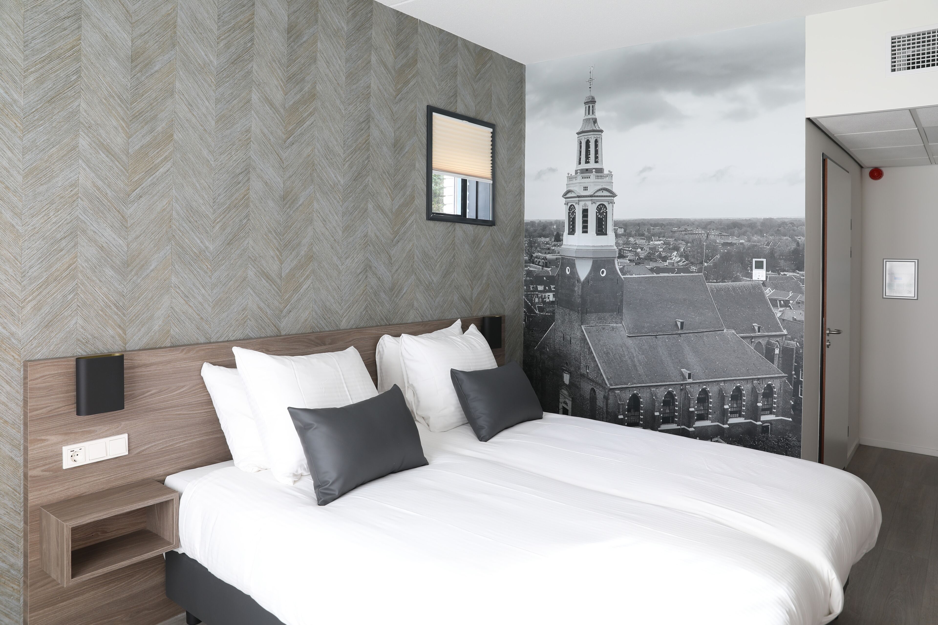 A modern hotel room with a large bed, a nightstand, a wall-mounted light, and a cityscape mural.