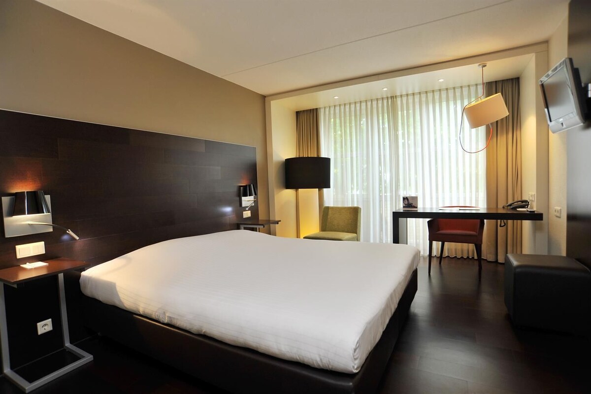 A hotel room with a large bed, a desk with a chair, a TV, and a lamp.