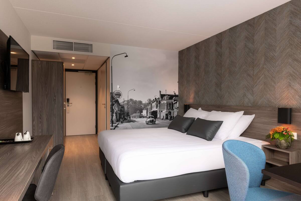 Een moderne hotelkamer met een groot bed, een bureau en een stoel. Er is een tv aan de muur en een tafeltje met een bloemenvaas.