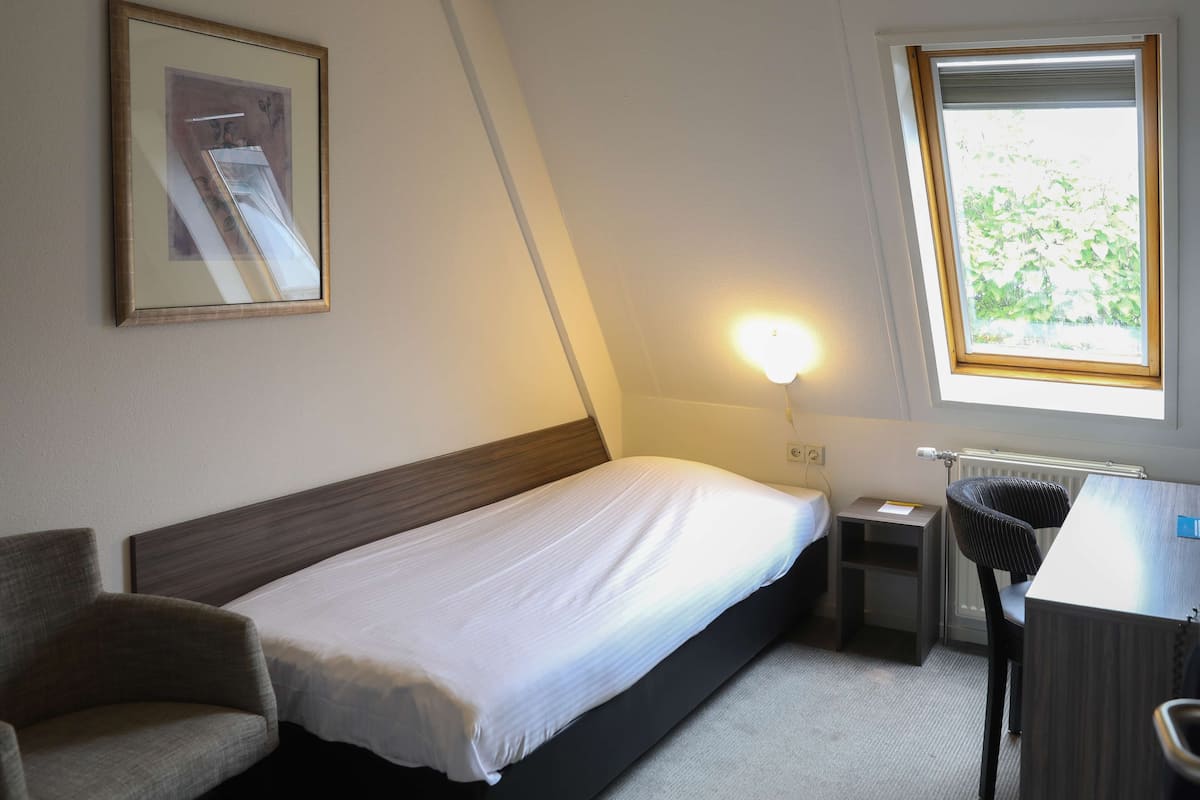 Een compact, verzorgd ingerichte slaapkamer met een enkel bed, een stoel, een bureau en een raam met uitzicht op groen.