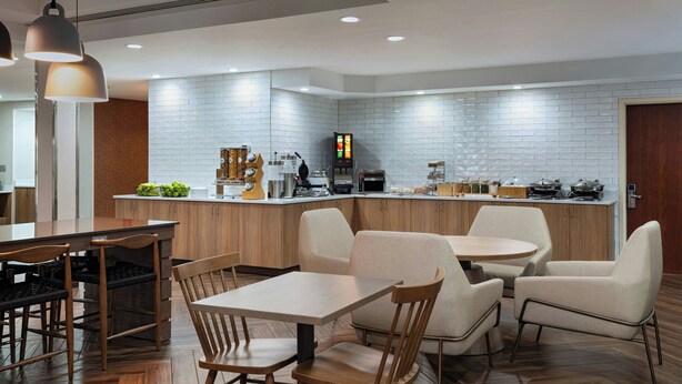 Een moderne keuken met een witte bakstenen spatwand, houten keukenkastjes en een centraal kookeiland met apparatuur. Er zijn twee eethoeken met tafels en stoelen.